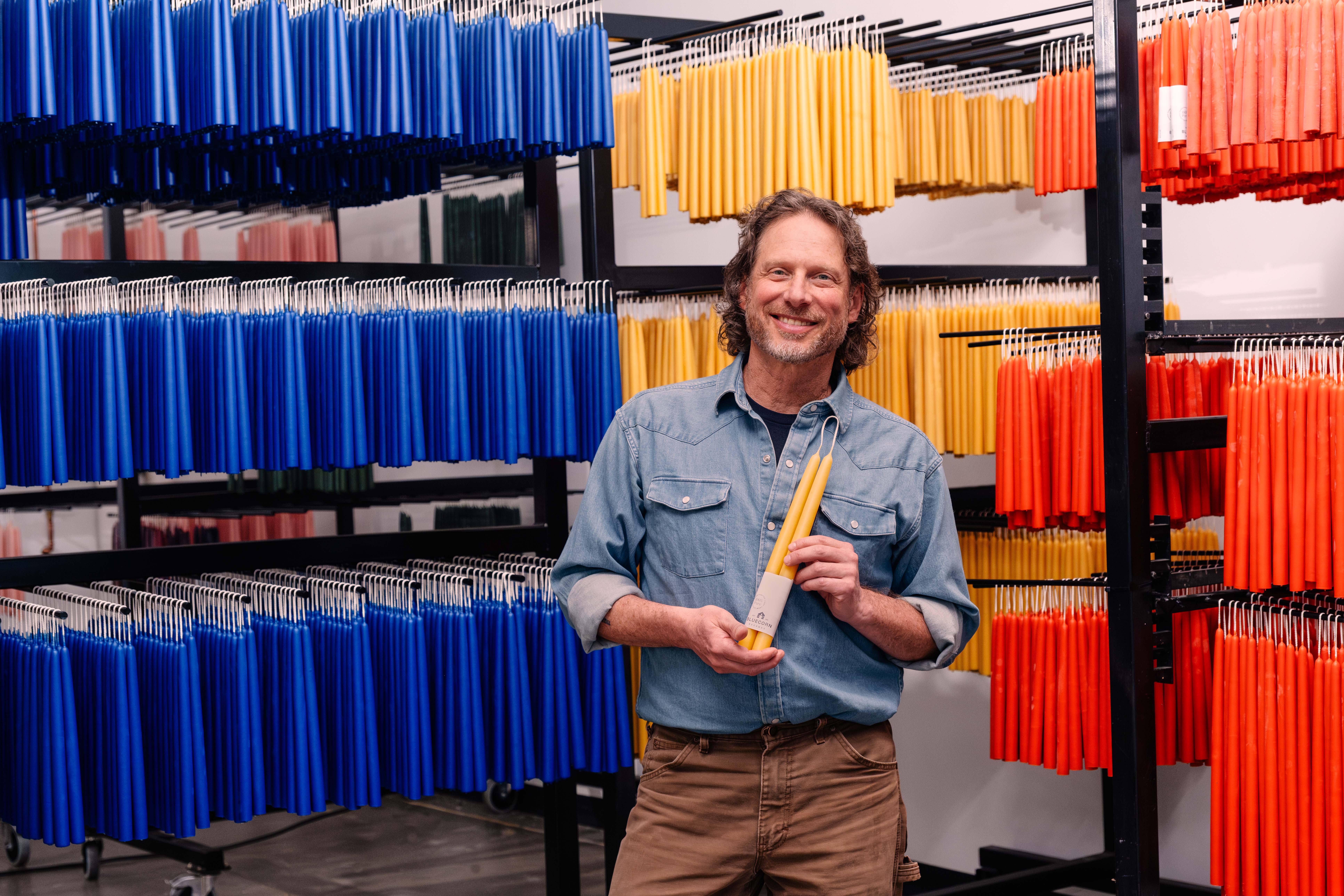 Jon Kornbluh stands in front of racks of colorful candlesticks, and holds a yellow one while smiling at the camera