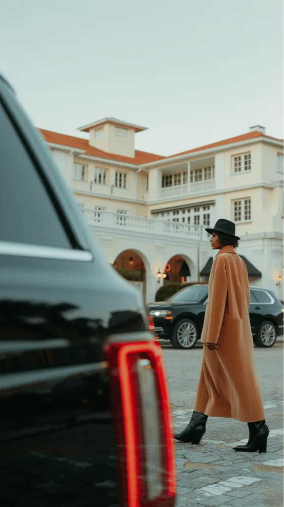 A woman in a brown coat walks past a black car.