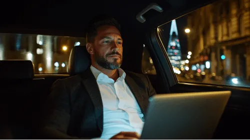 A man in a suit sitting in a car and using a laptop.
