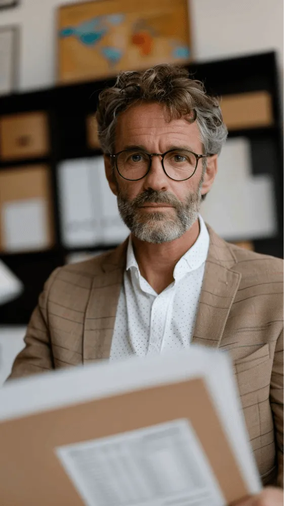 A man with a beard and glasses sitting in front of a stack of papers.