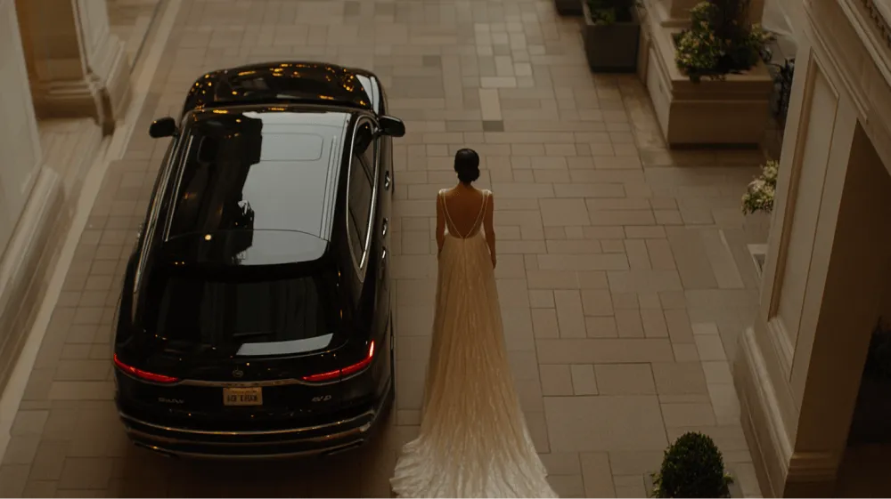 A woman in a white dress stands in front of a black car.