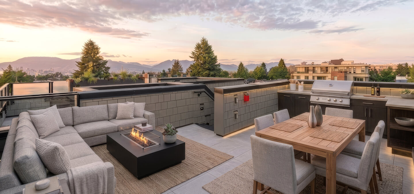 Modern rooftop balcony with gray sectional sofa, fire pit, wooden dining table, and outdoor grill at sunset.