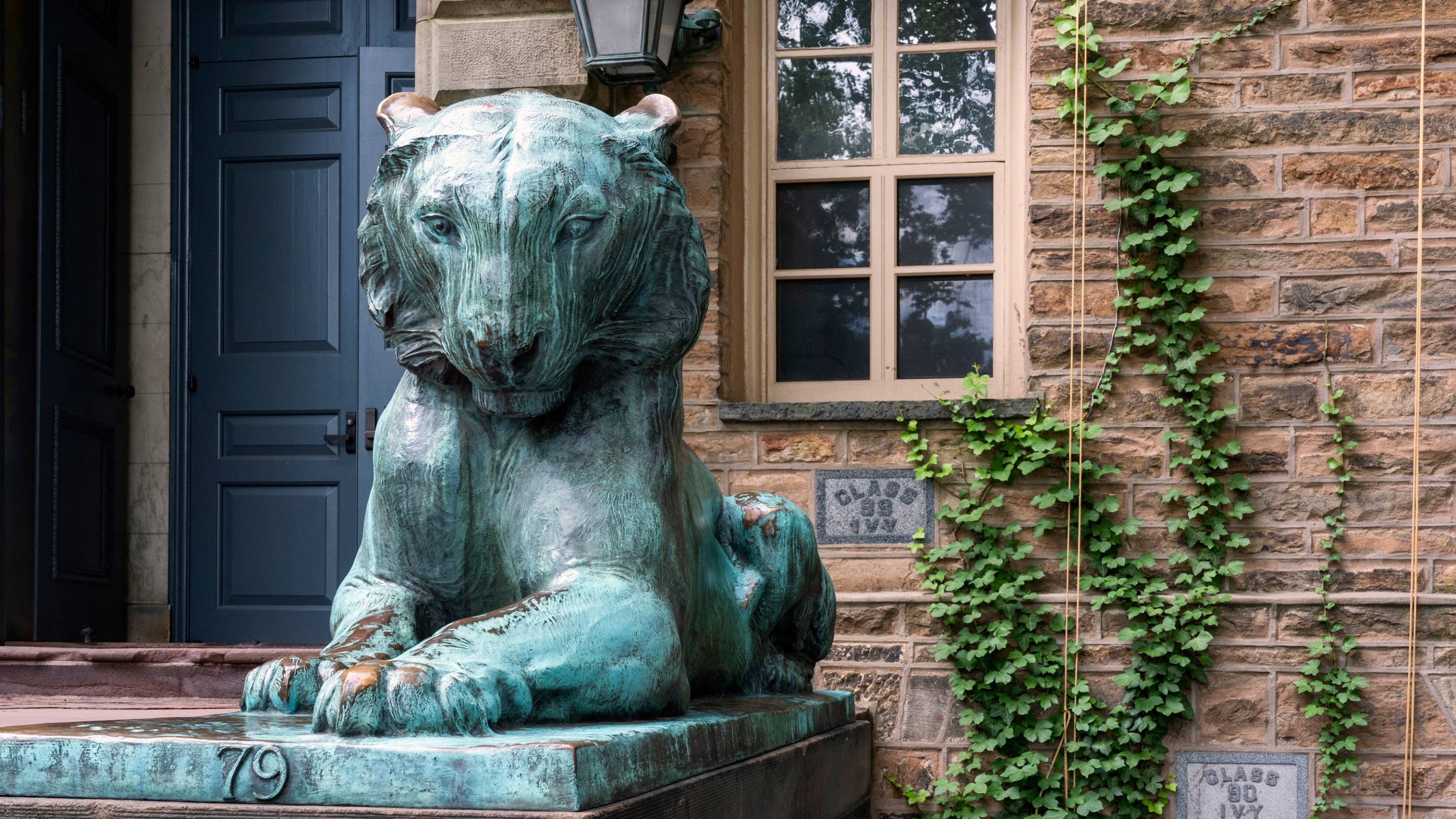 Bronze tiger statue with a green patina sits outside a stone building entrance, with a door, window, and climbing ivy in the background.