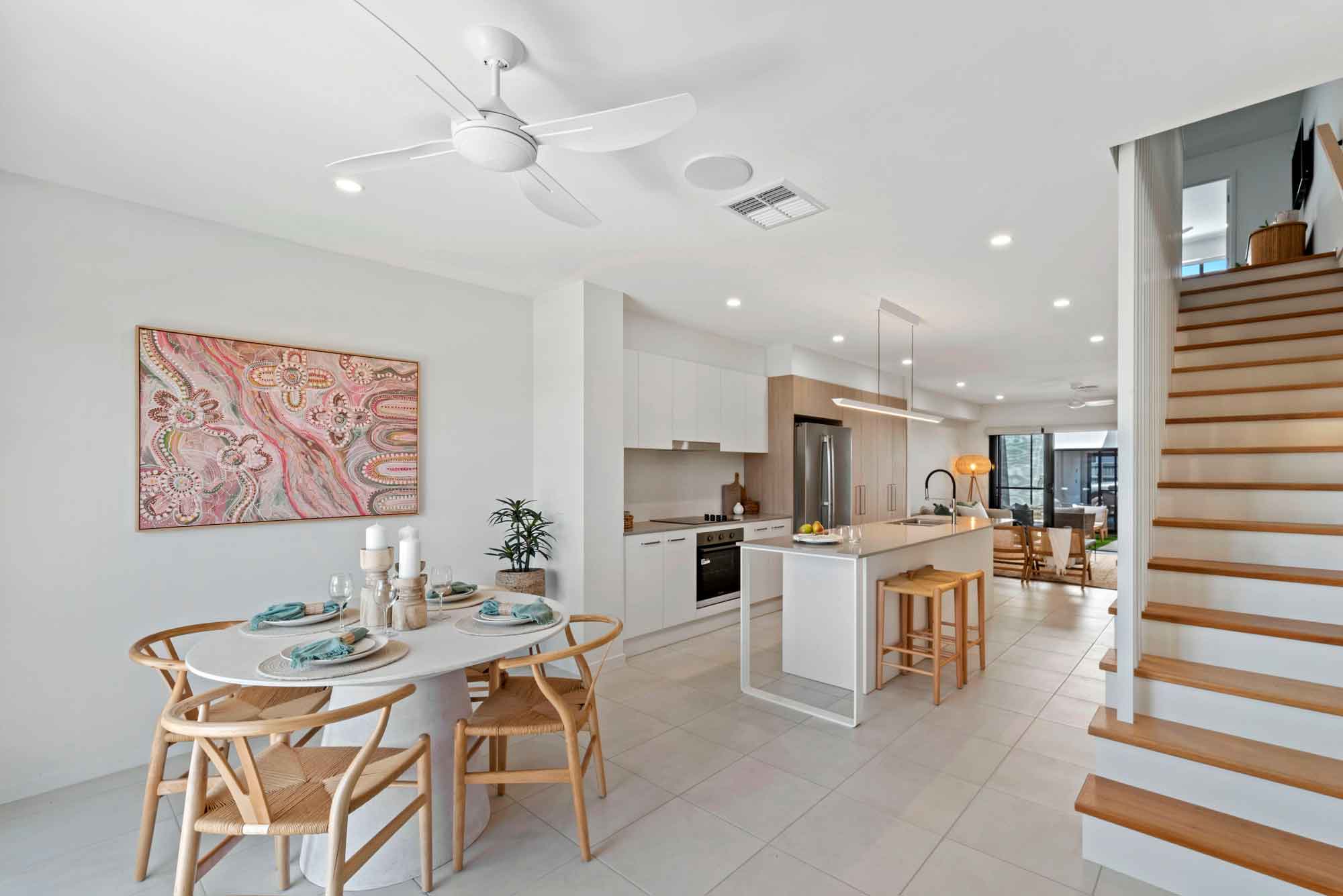 Open-plan kitchen and dining area featuring white cabinetry, timber accents, stone island bench with breakfast seating, stainless steel appliances and round dining table in a modern coastal home.