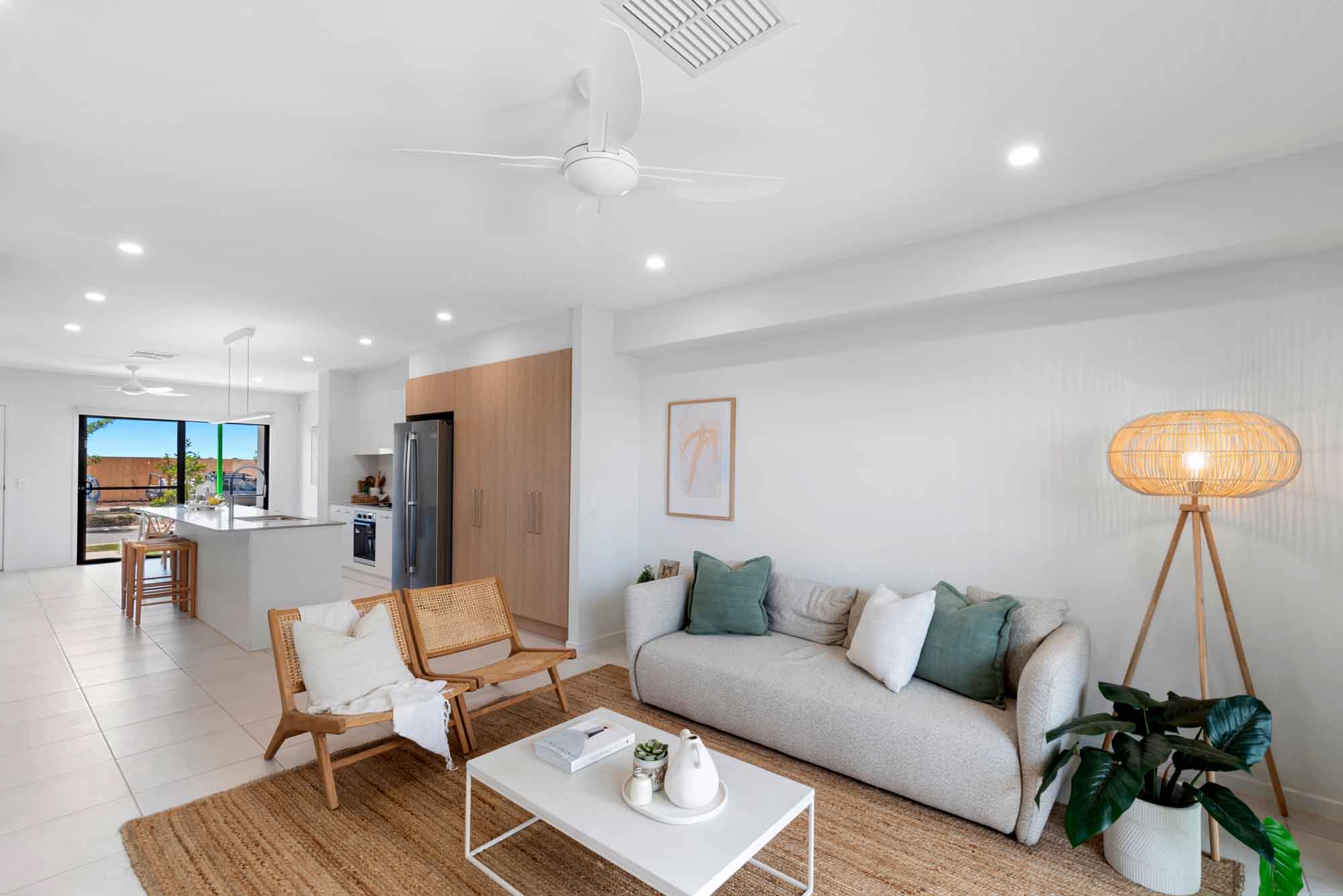 Bright open-plan living room with neutral sofa, timber accent chairs, woven rug, floor lamp and seamless flow to a modern kitchen in a contemporary terrace-style home.