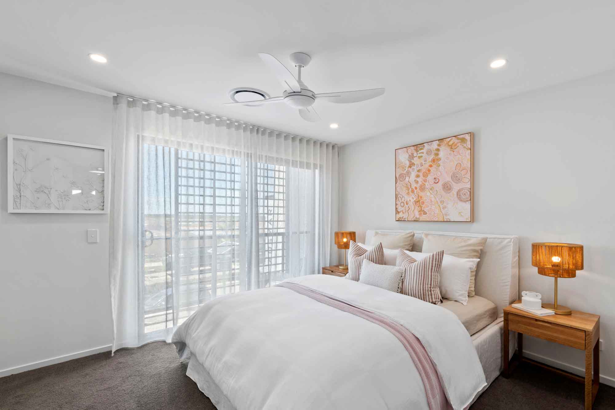 Light-filled master bedroom with sheer floor-to-ceiling curtains, soft neutral styling, timber bedside tables, warm ambient lighting, ceiling fan and contemporary artwork in a modern Sunshine Coast home.