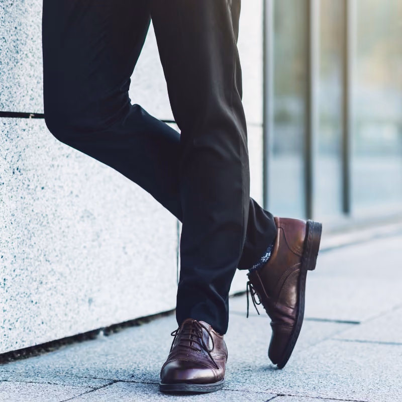Beine einer Person in dunkler Hose und Lederschuhen, lässig an eine Wand gelehnt