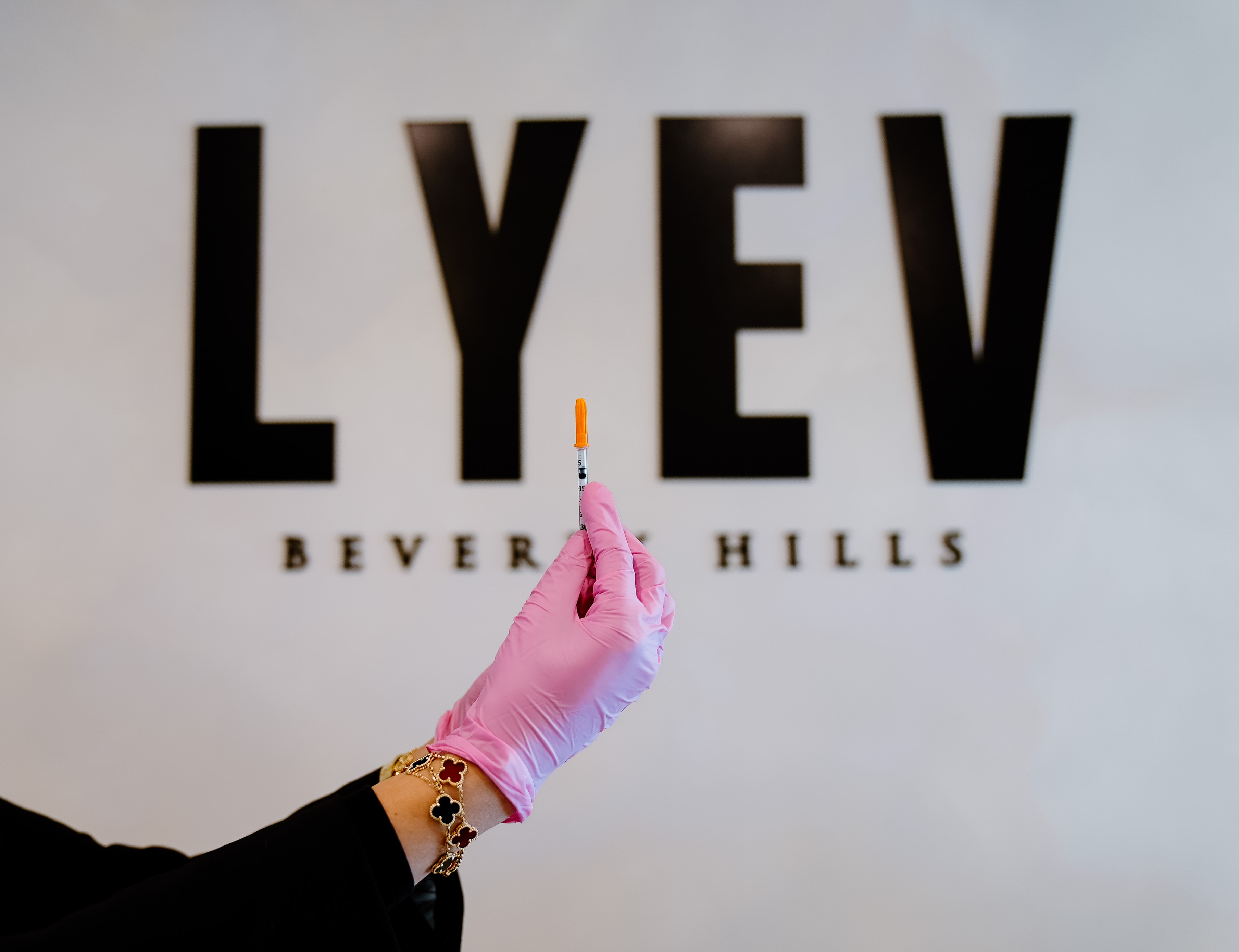 A pink-gloved hand holds a capped syringe in front of the LYEV Beverly Hills sign.