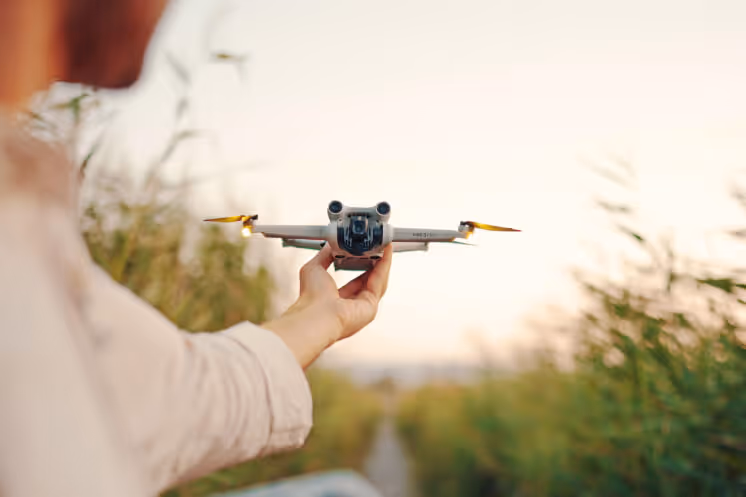 Person holding a small drone with yellow propellers outdoors near greenery at sunset.