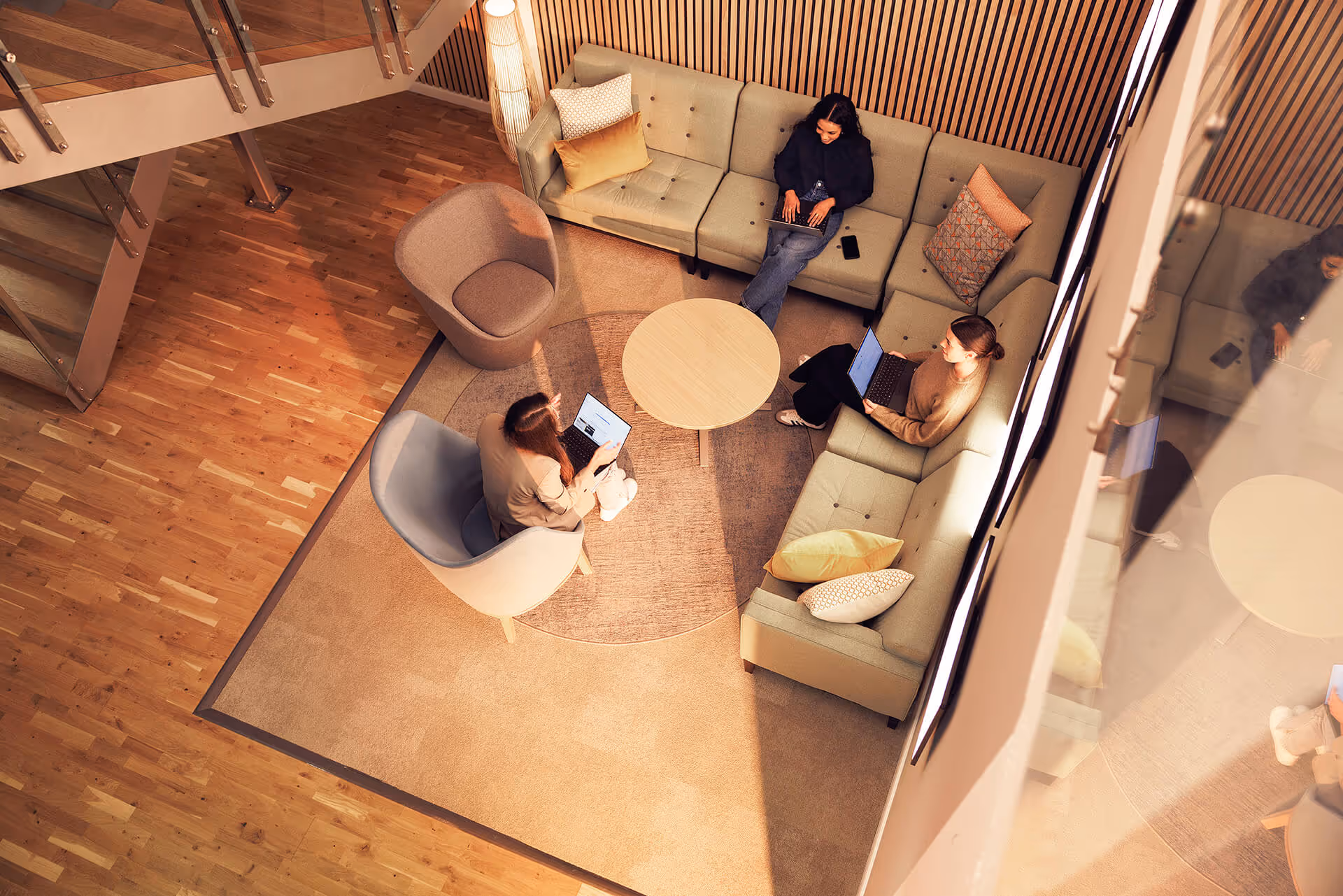 Three people working on laptops seated around a round wooden table in a modern office lounge with a sectional sofa and armchairs.