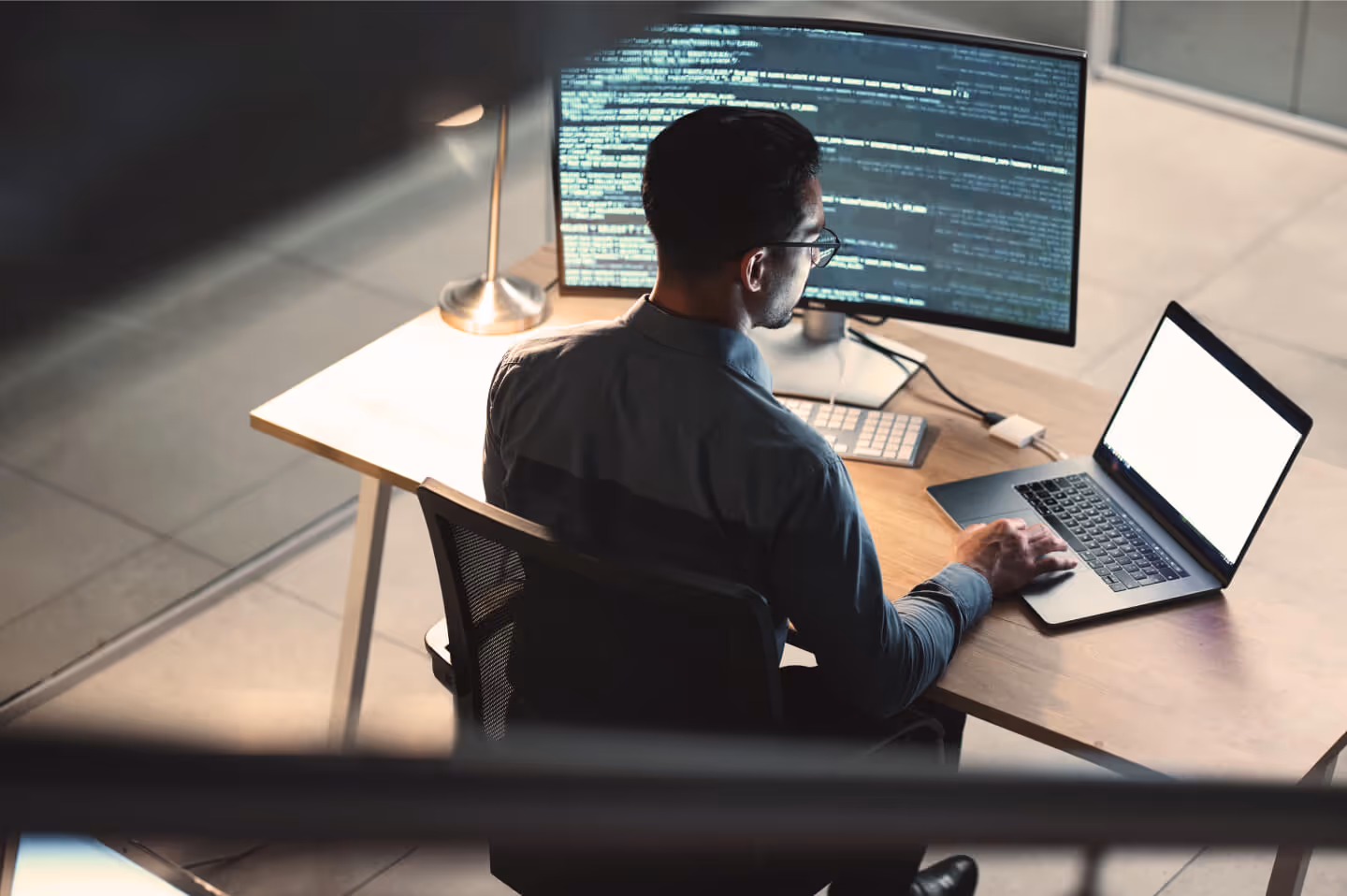 Man wearing glasses working at a desk using a laptop and a curved monitor displaying lines of code.