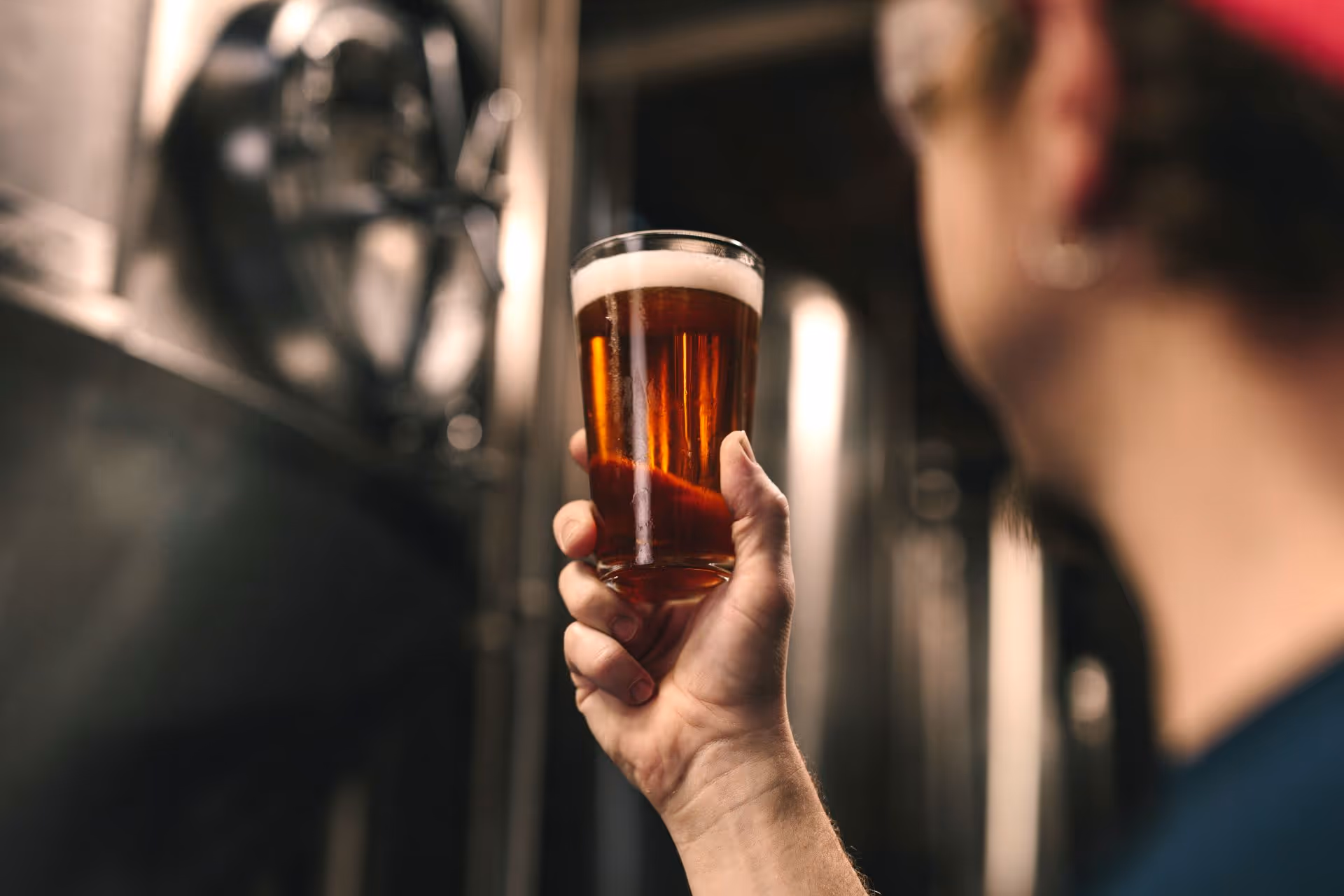 Person holding up a glass of amber beer with foam on top in a brewery setting.