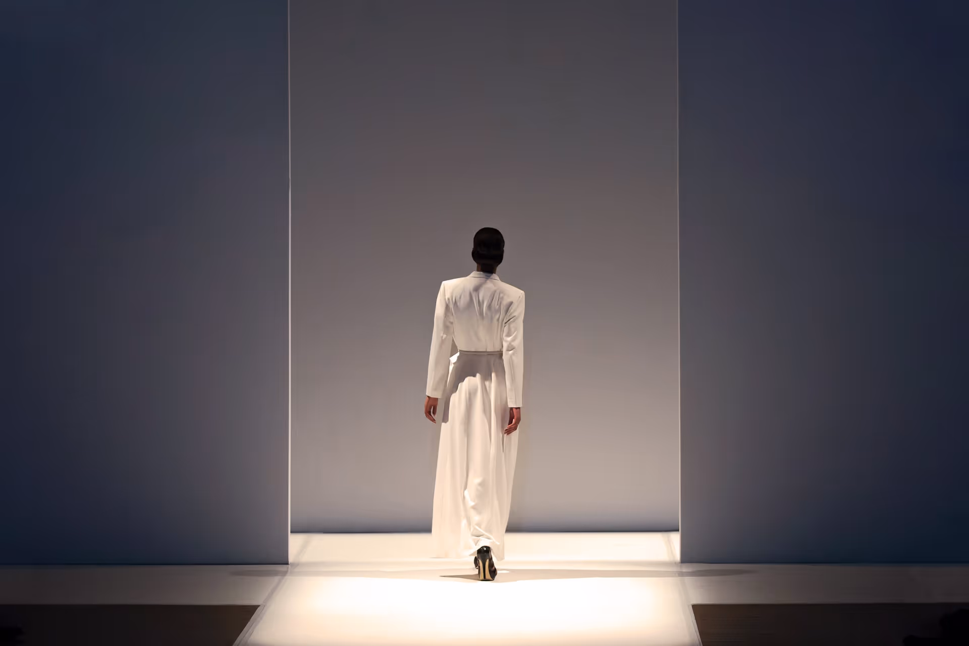 Model walking away on a runway wearing a long white dress with long sleeves in a minimalist setting.