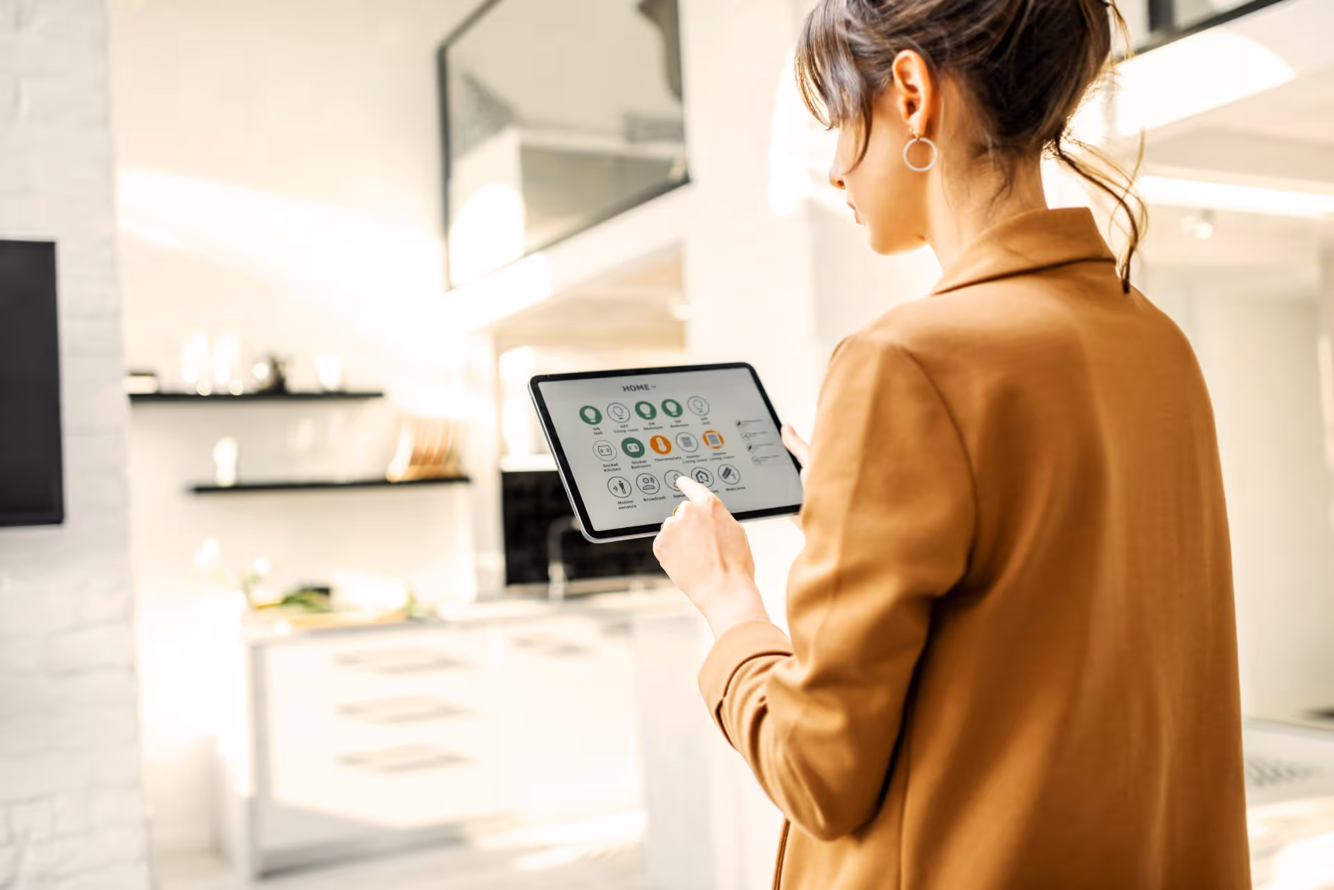 Woman in a brown coat using a tablet to control smart home devices in a bright modern kitchen.