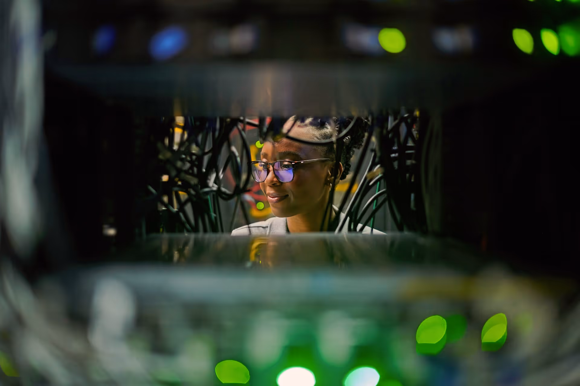 Technician with glasses working behind server cables illuminated by green and blue lights.