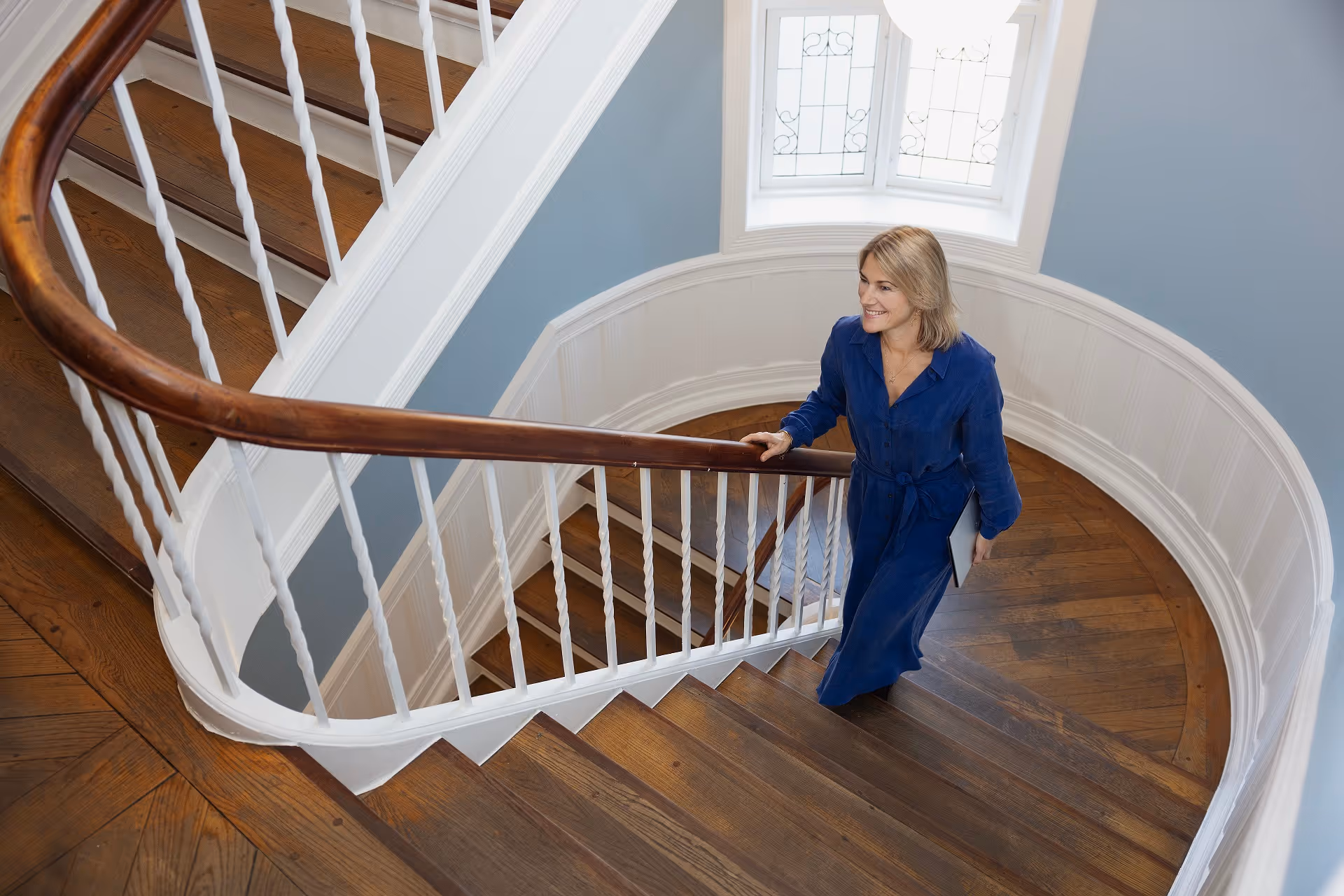 Smiling woman in blue dress ascending a curved wooden staircase with white banisters in a bright interior.