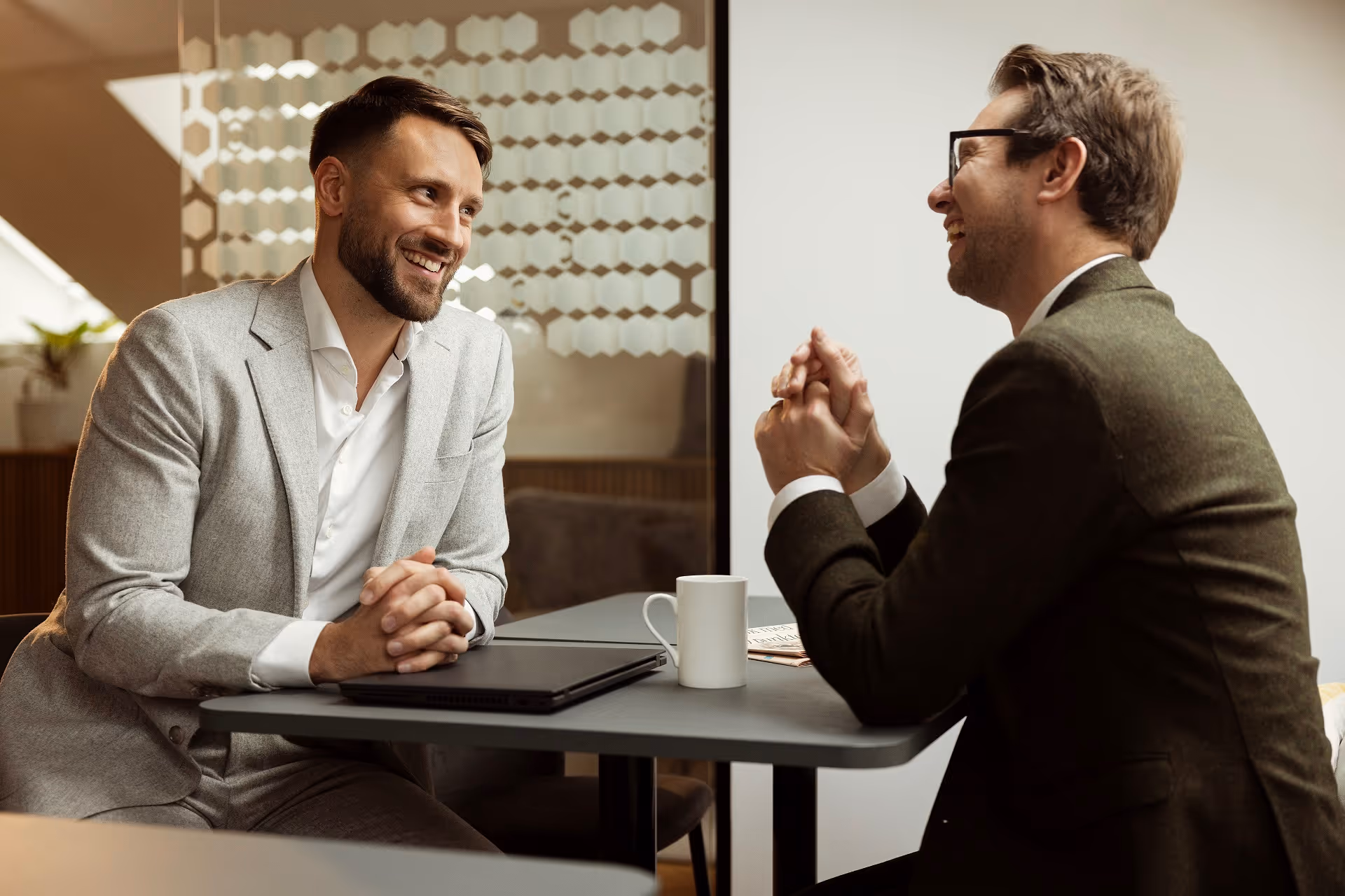 Two businessmen smiling and talking across a table in a modern office setting.