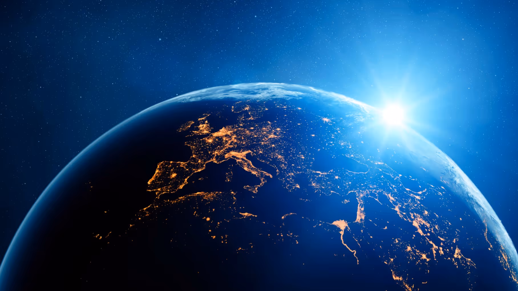 View of Earth from space at night showing illuminated cities across Europe with the sun rising over the horizon.