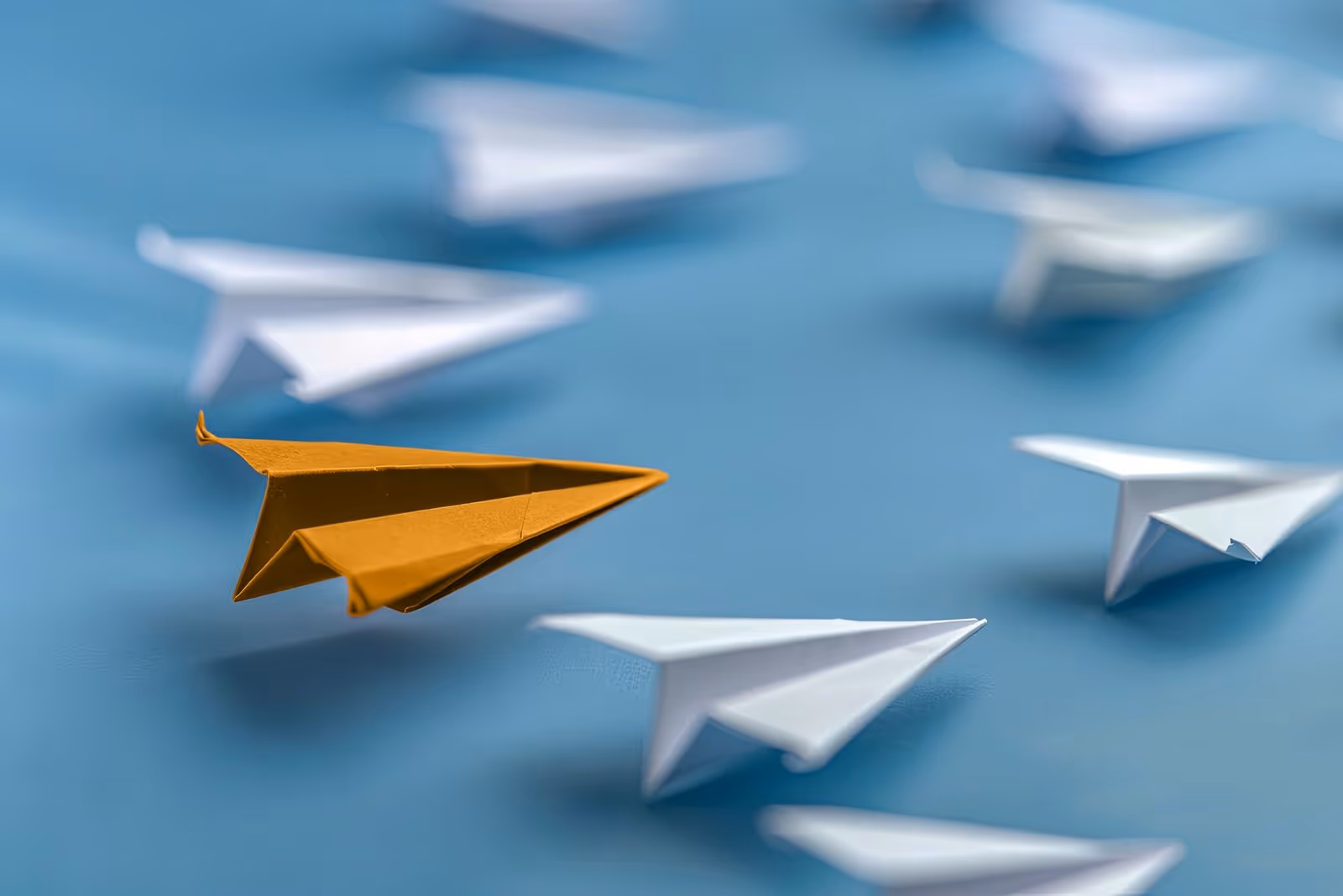 Orange paper airplane leading a group of white paper airplanes on a blue surface.