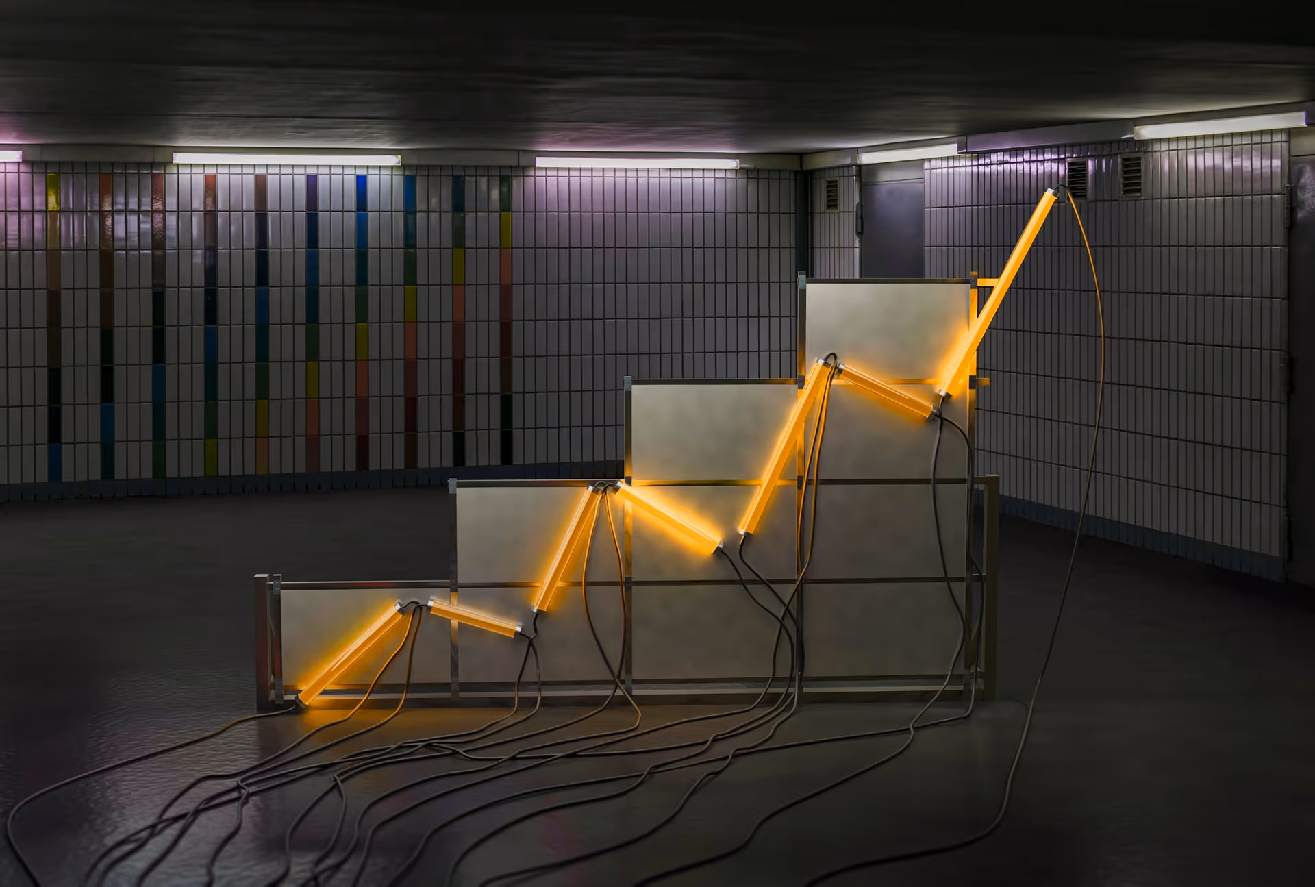 Neon yellow light tubes arranged in a rising zigzag pattern on a metal frame, set in a tiled underground corridor.