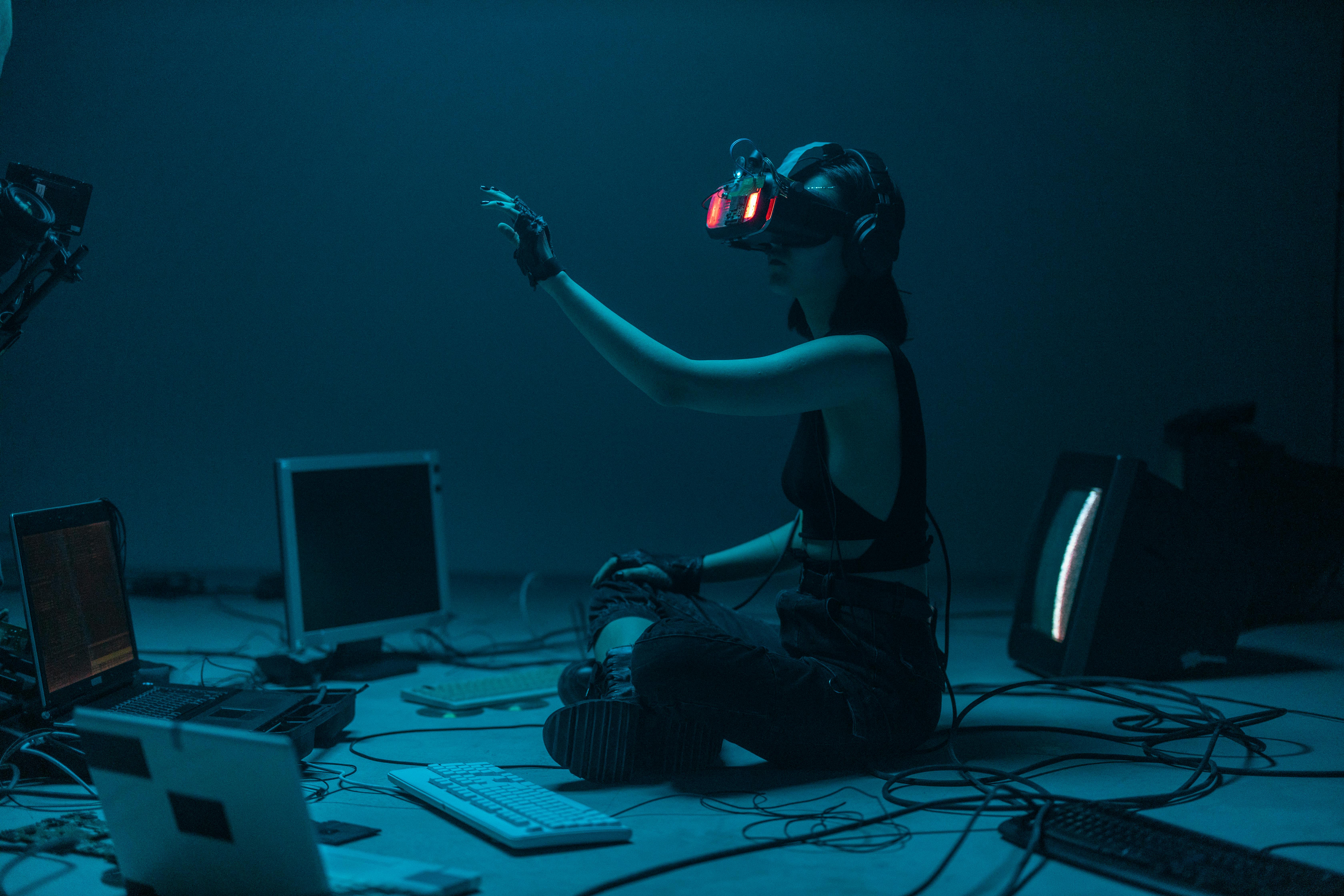 Person sitting cross-legged on the floor in a dark room, wearing a VR headset and headphones, surrounded by old computer monitors, keyboards, and tangled wires, reaching out with a gloved hand as if interacting with a virtual environment.