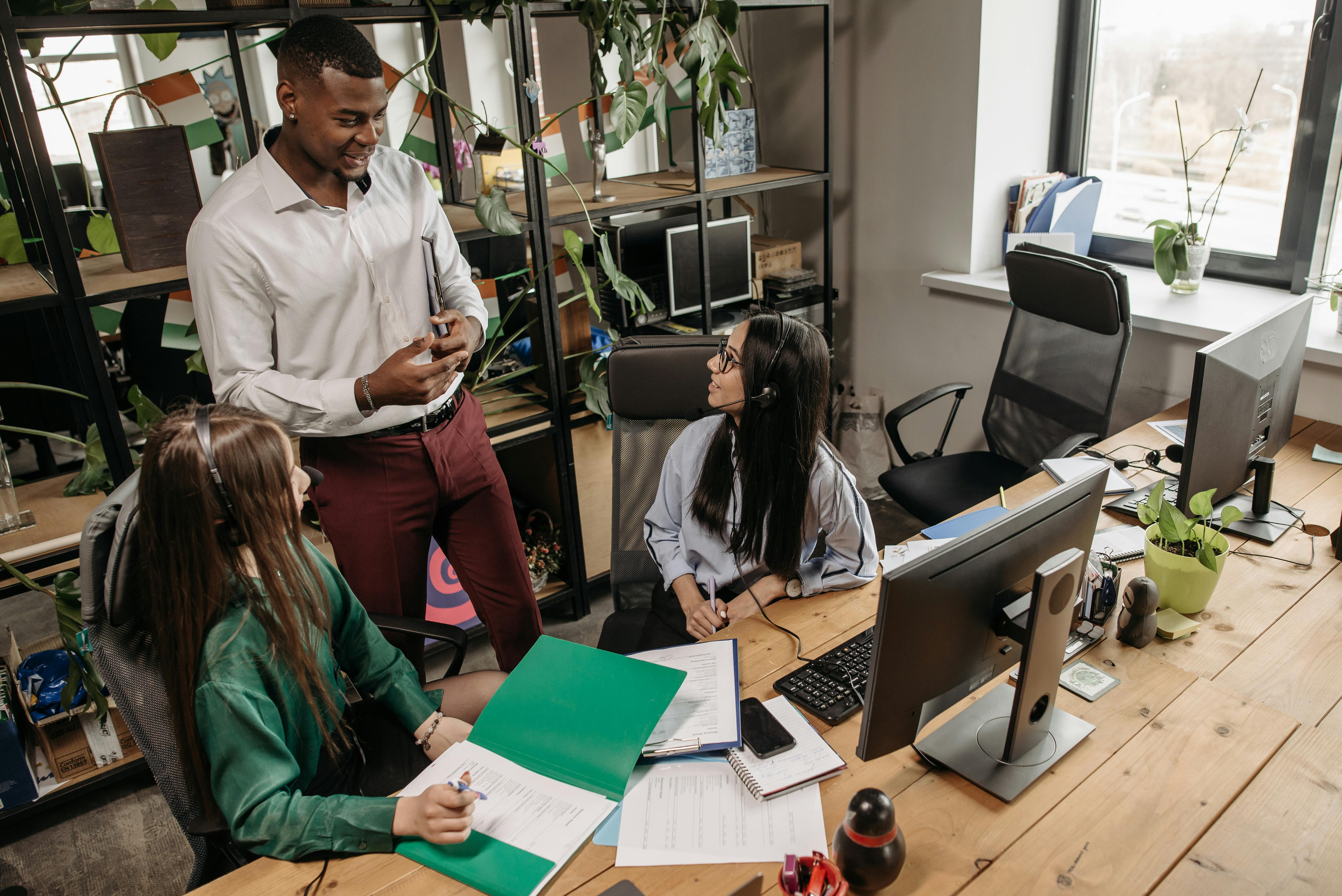 Three colleagues having a collaborative discussion in a modern office setting with computers, documents, and indoor plants around them.