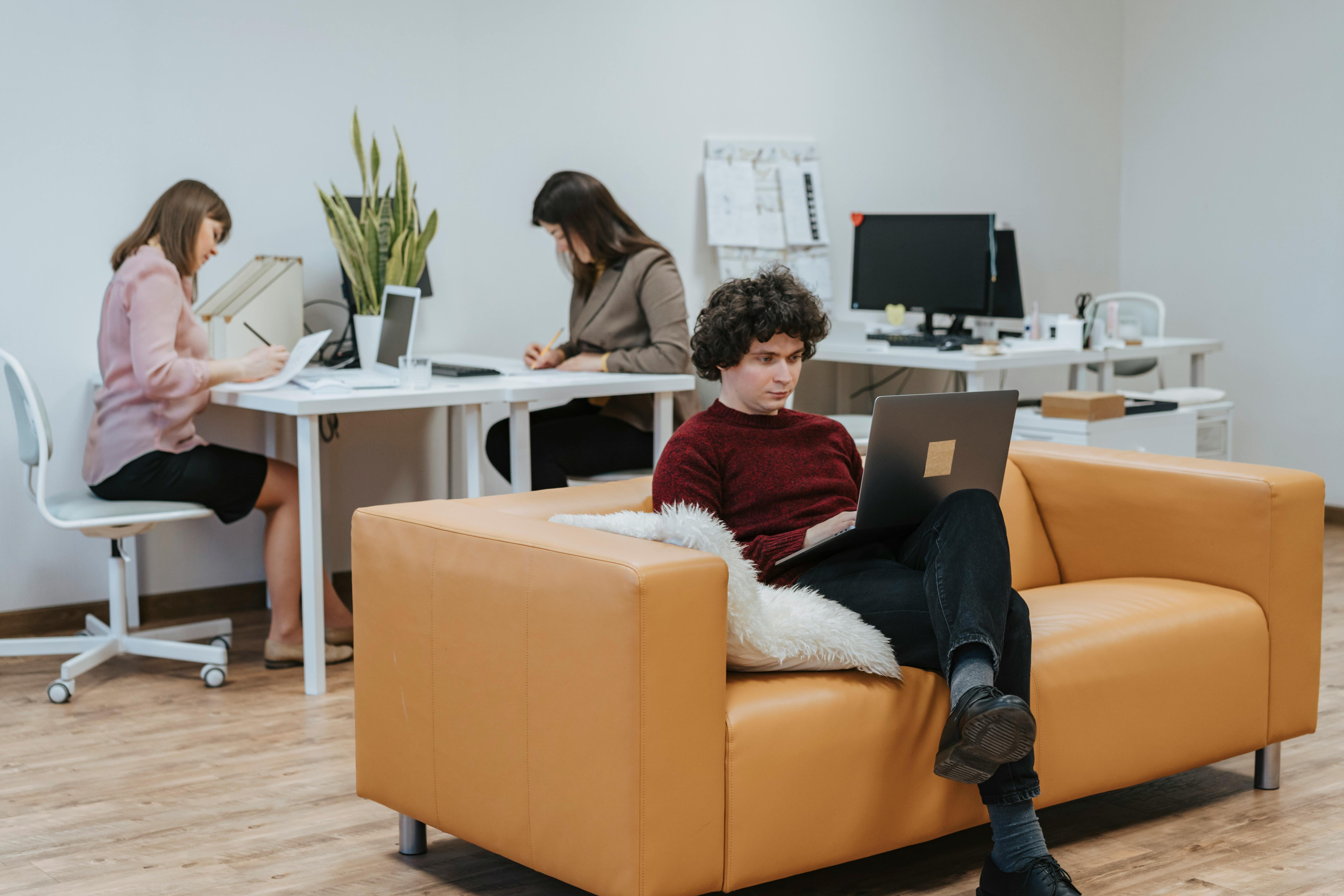 Man working on a laptop while sitting on a tan sofa, with two women focused on paperwork and computers at desks in a modern office setting.