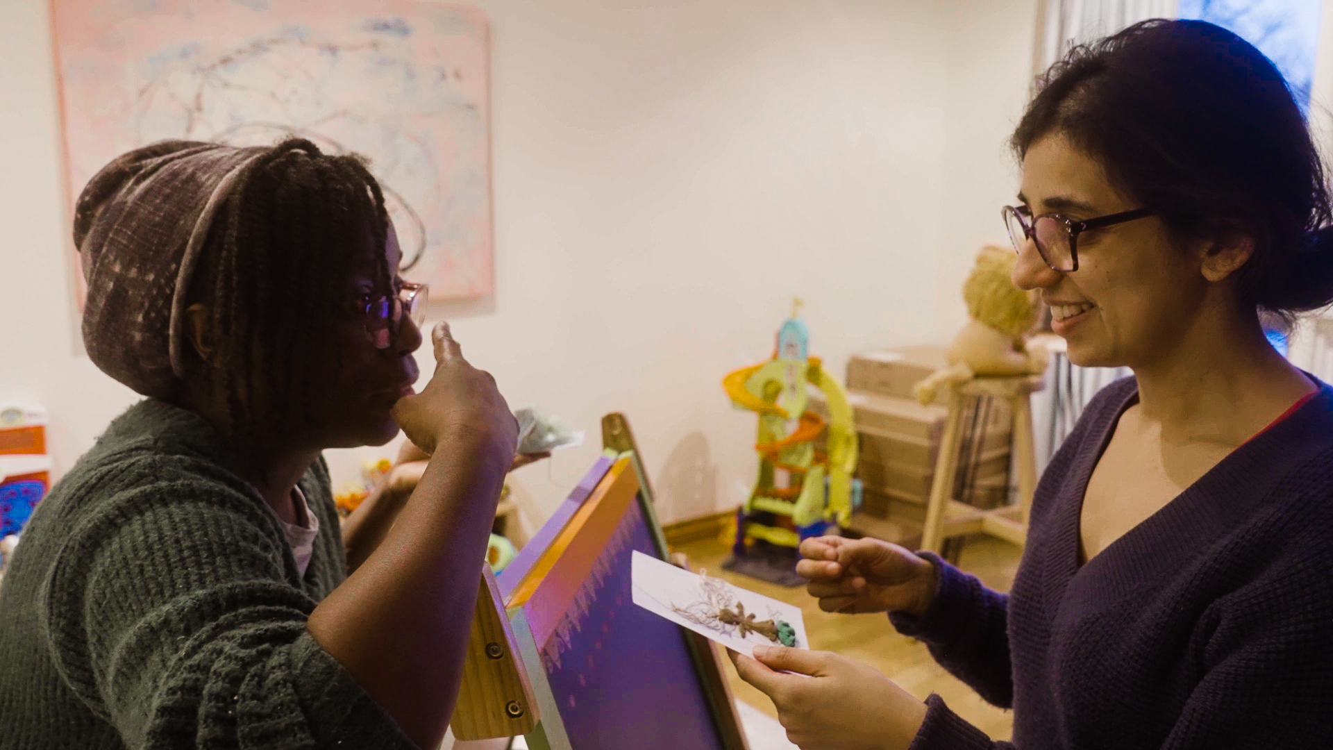 Jenelle and Kanika stand facing each other in a room of bright toys. Kanika holds a piece of paper and a small clay figure, Jenelle is pointing to herself.
