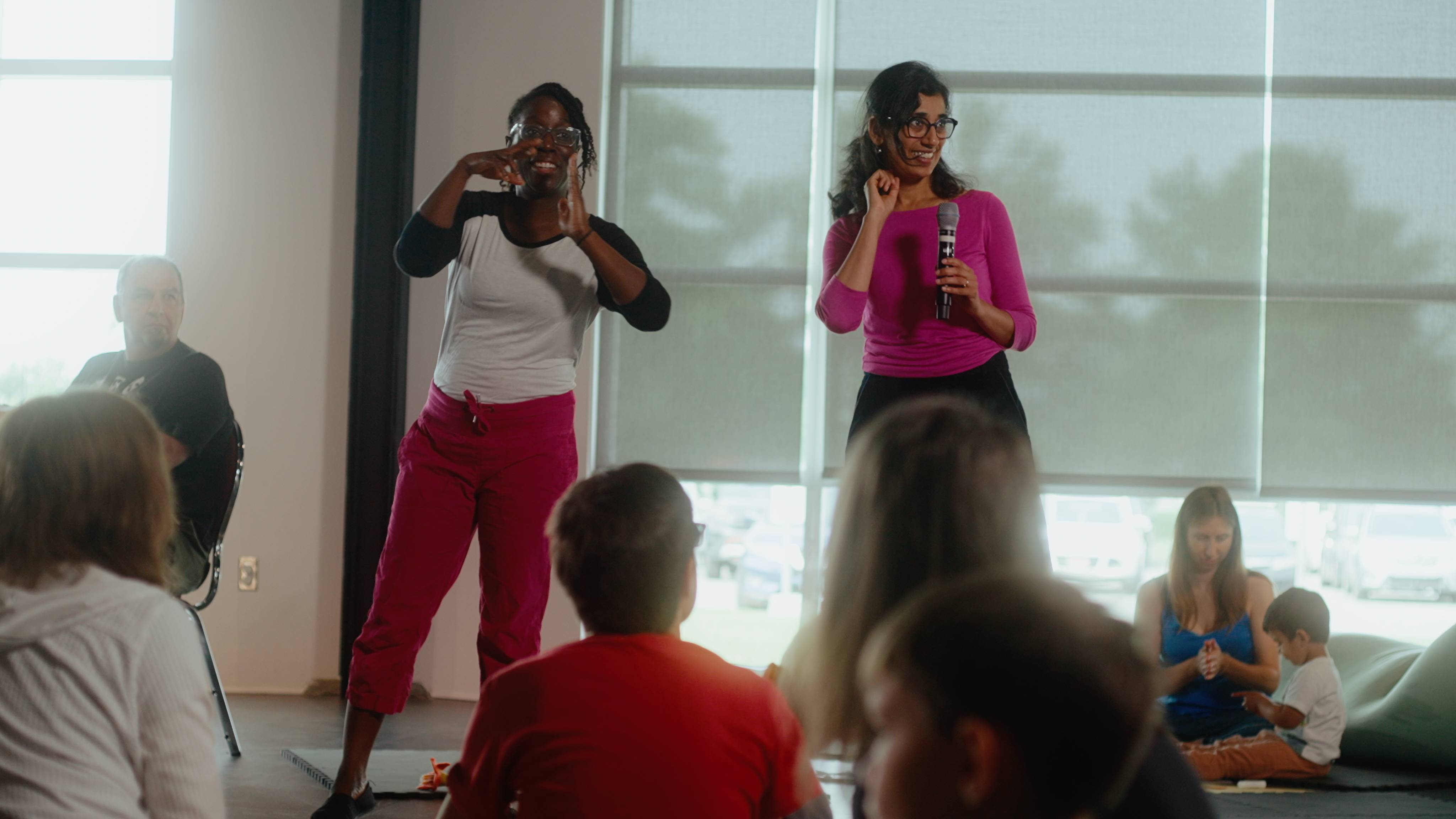 Jenelle and Kanika on stage presenting in front of a few people. Both women are smiling as Jenelle gestures, mid-sign, while Kanika holds a microphone.