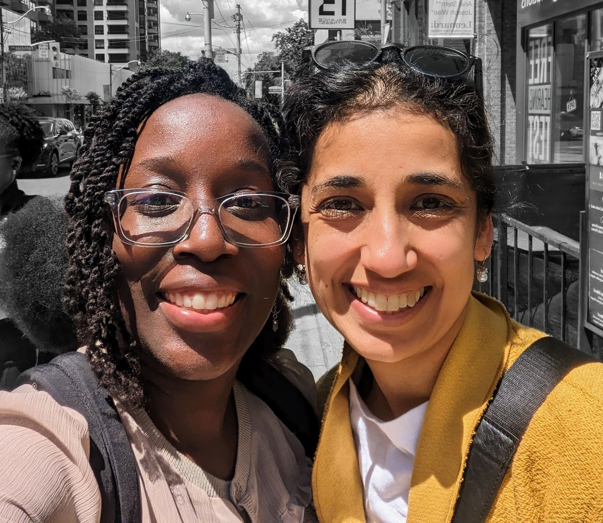A selfie of Jenelle and Kanika smiling with their faces close together, the women are in colour while the background of a city street is in black and white.