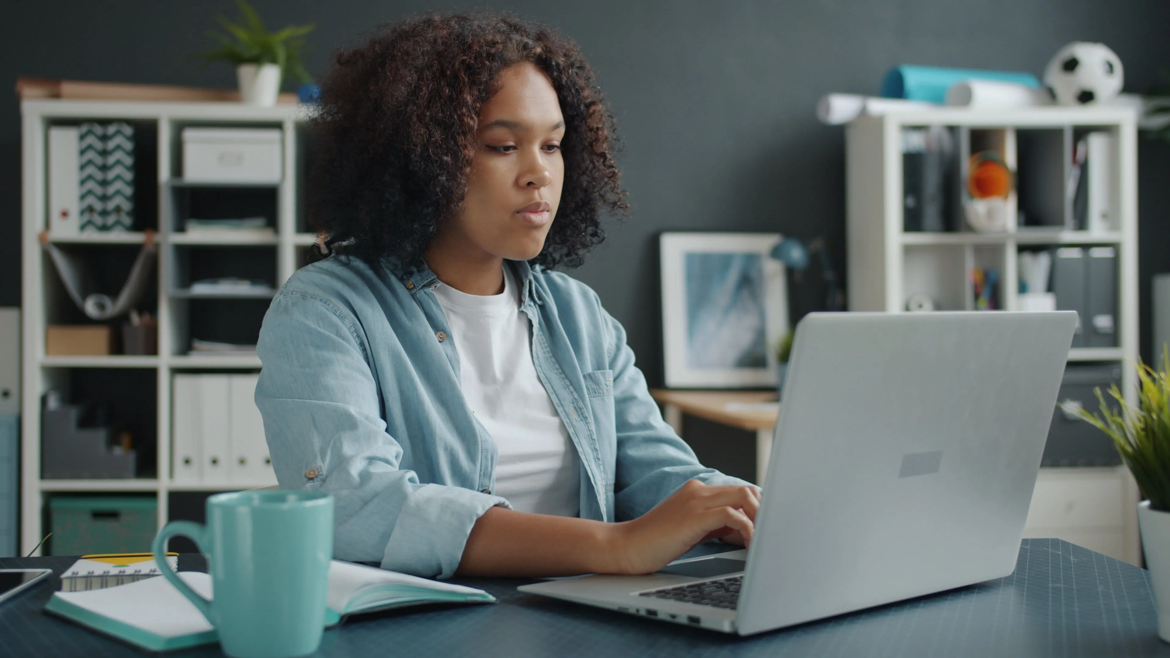Woman in denim shirt working on a laptop at a desk with a mug and open notebook.