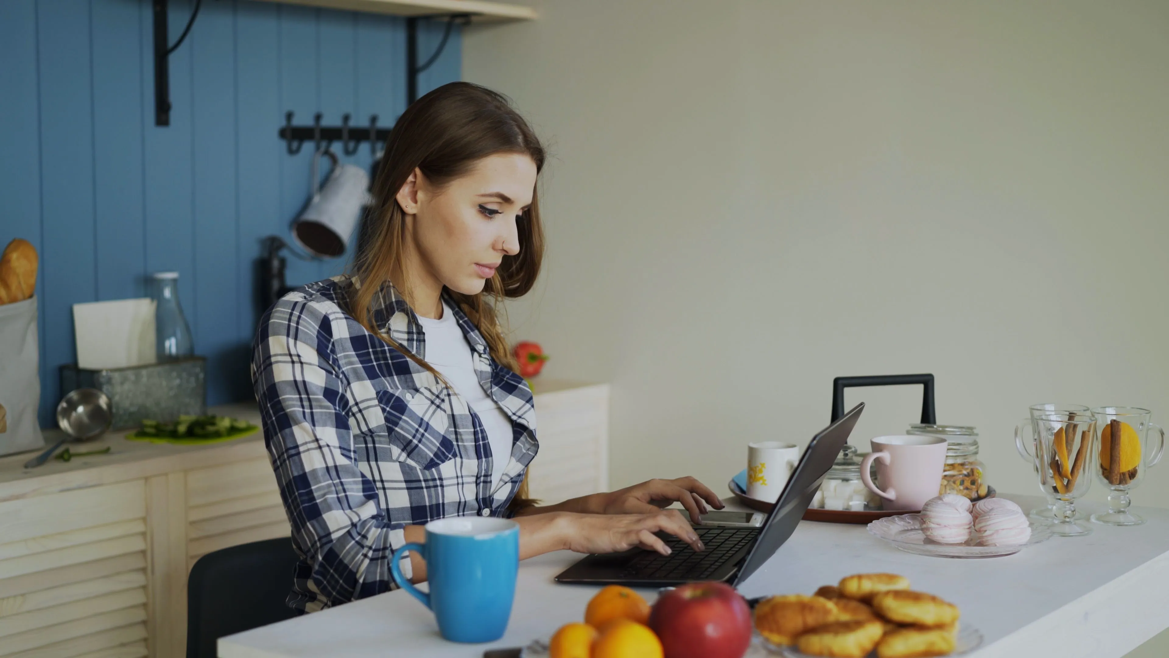Woman in plaid shirt typing on laptop at kitchen table with fruit, pastries, and mugs nearby.