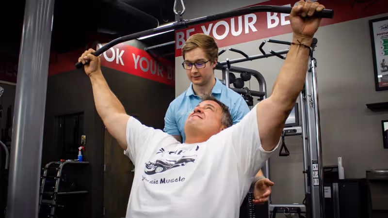 A man performs a lat pulldown exercise on a machine while another man assists him at a gym.