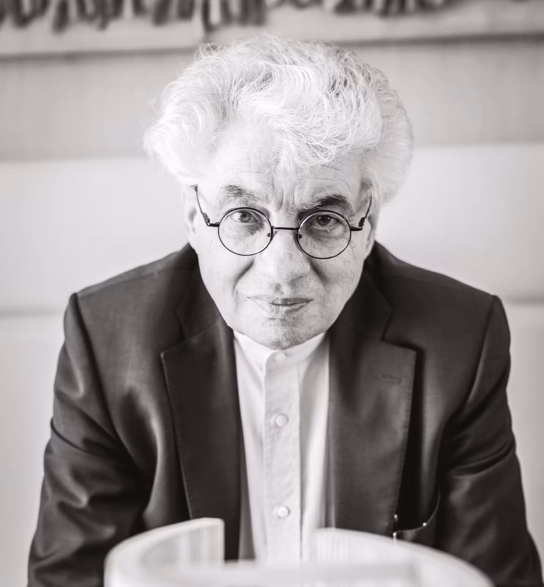 Mario Botta.
Retrato en blanco y negro de un hombre mayor con cabello canoso y rizado, usando gafas redondas y un saco oscuro, mirando hacia la cámara.