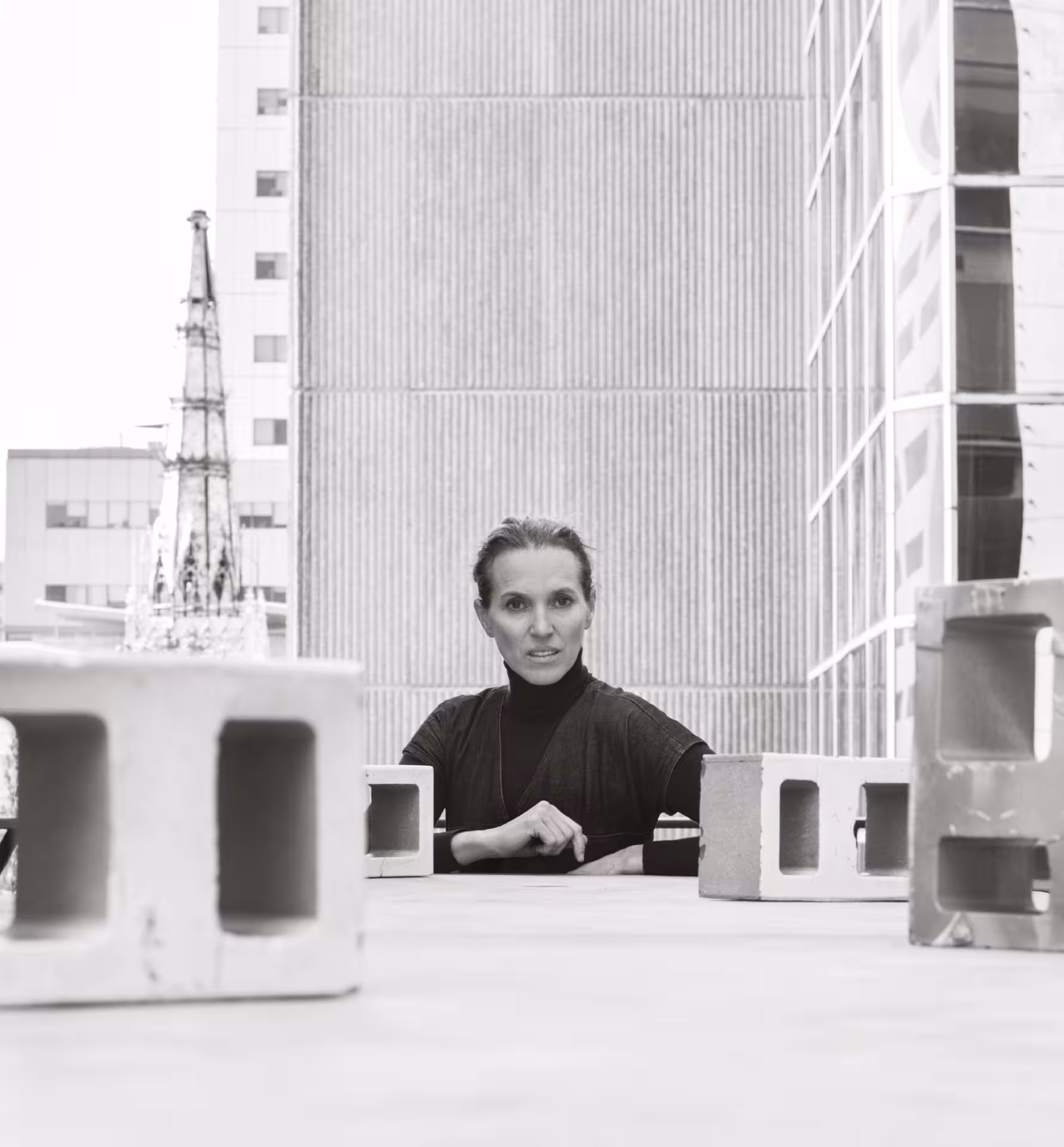 Tatiana Bilbao.
Mujer de cabello recogido y suéter oscuro sentada entre bloques de concreto en una azotea urbana con edificios altos al fondo.
