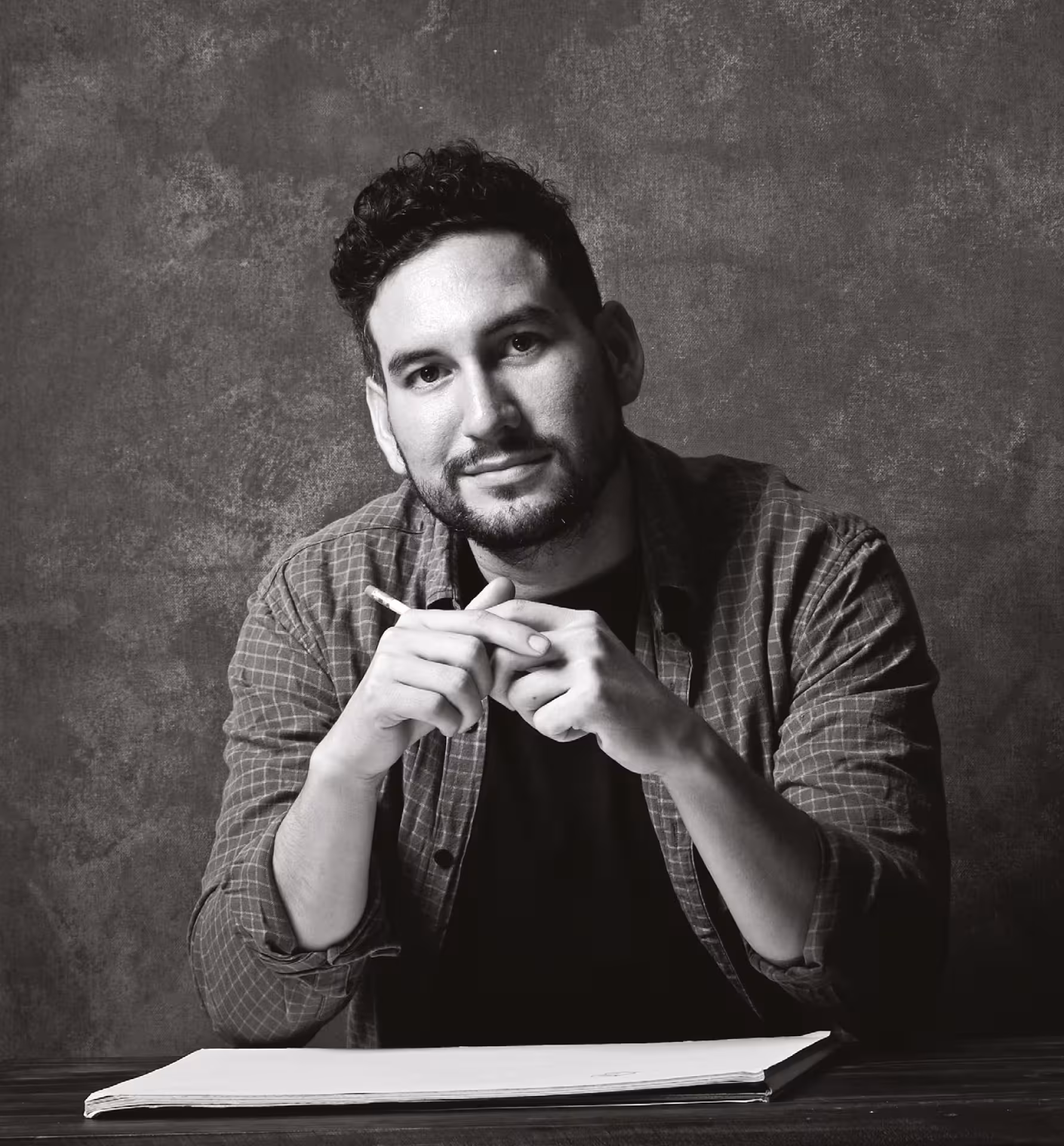 Natura Futura.
Hombre sentado frente a una mesa con un cuaderno abierto, sosteniendo un lápiz y mirando a la cámara en un retrato en blanco y negro.