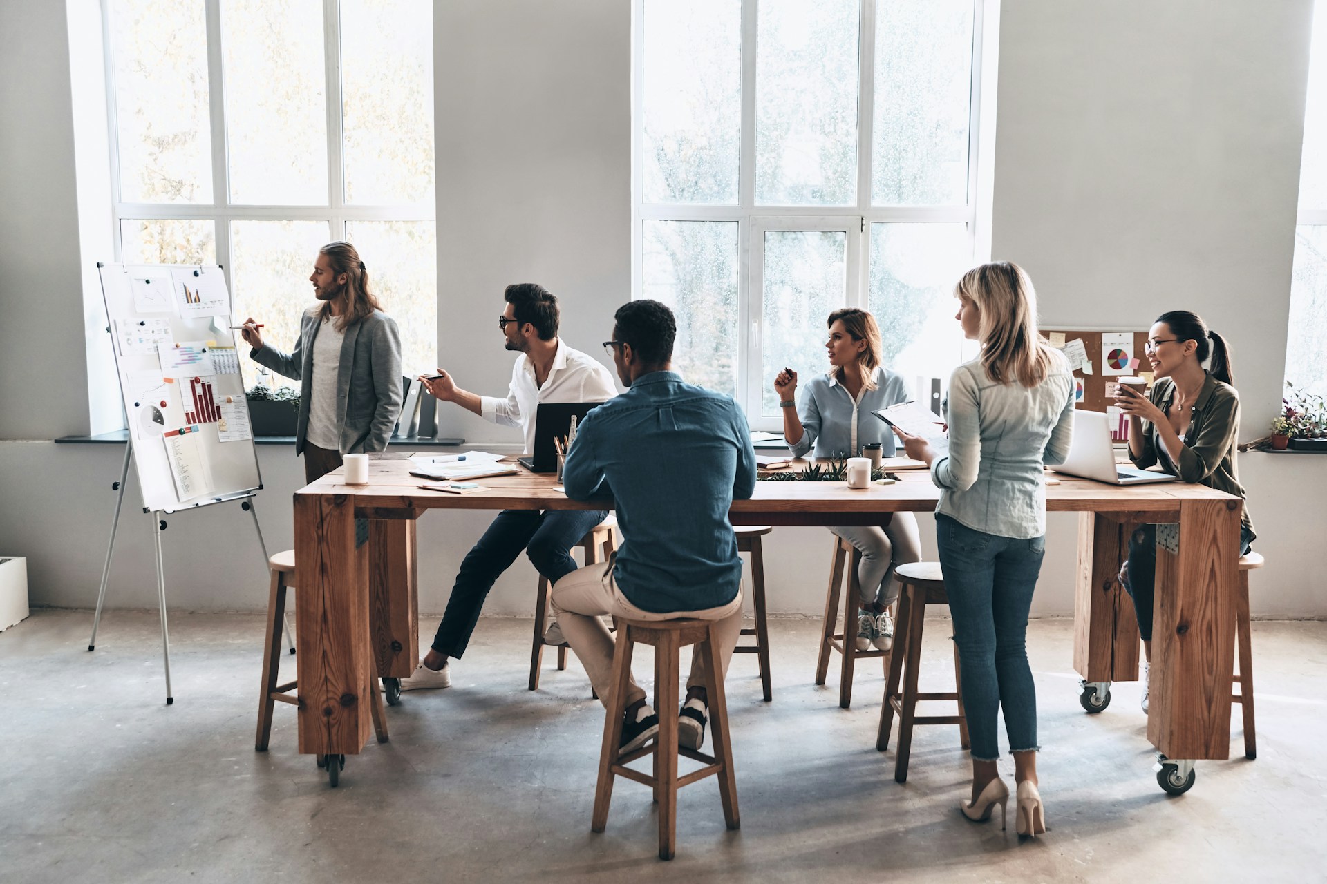 Personen bei einem Teammeeting, die Präsentationen diskutieren und Ideen austauschen. Büroatmosphäre mit einem langen Tisch und großen Fenstern.