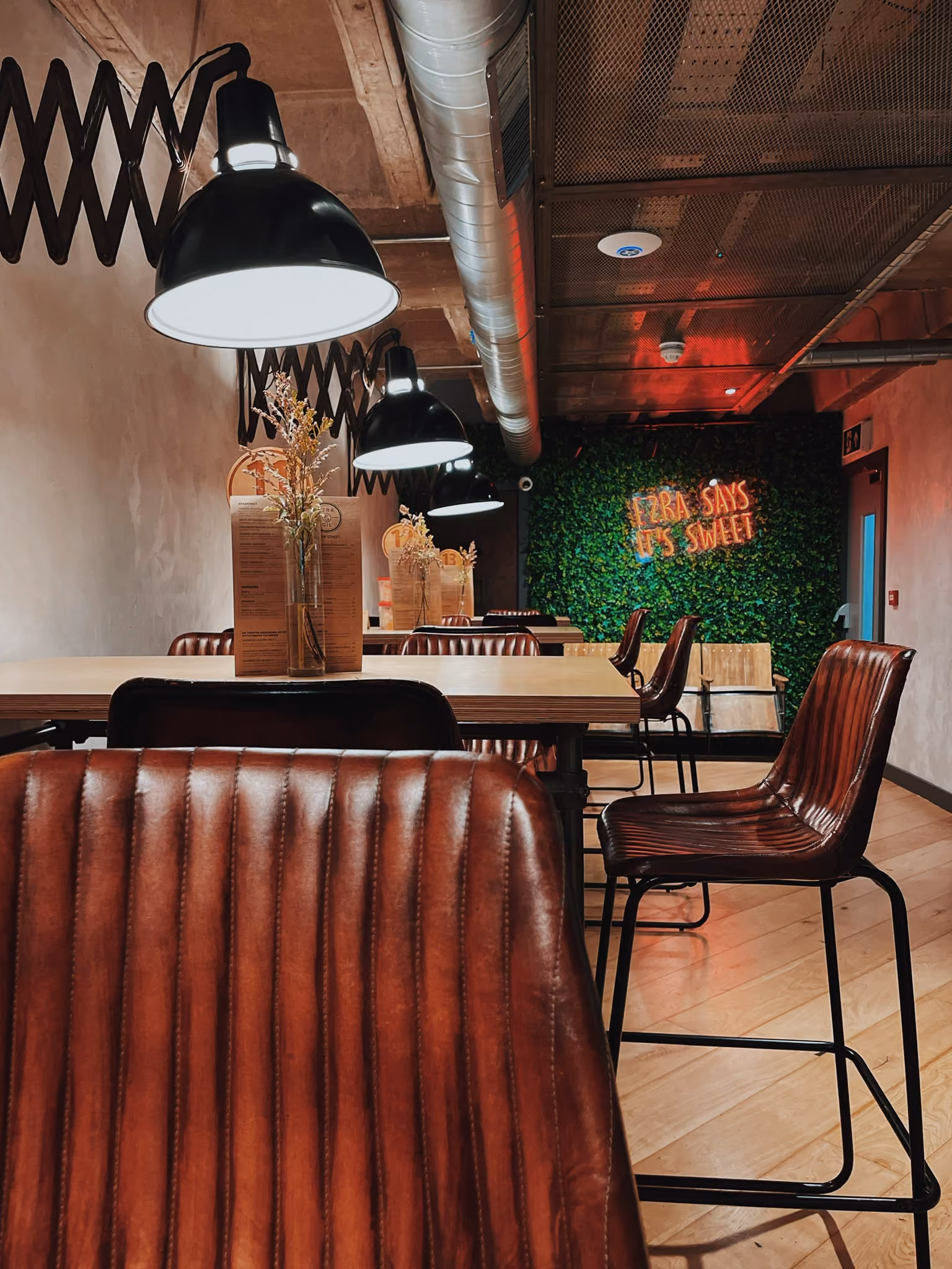 Leather bar stools and pendant lamps beside a living wall with neon sign at Ezra & Gil Peter Street
