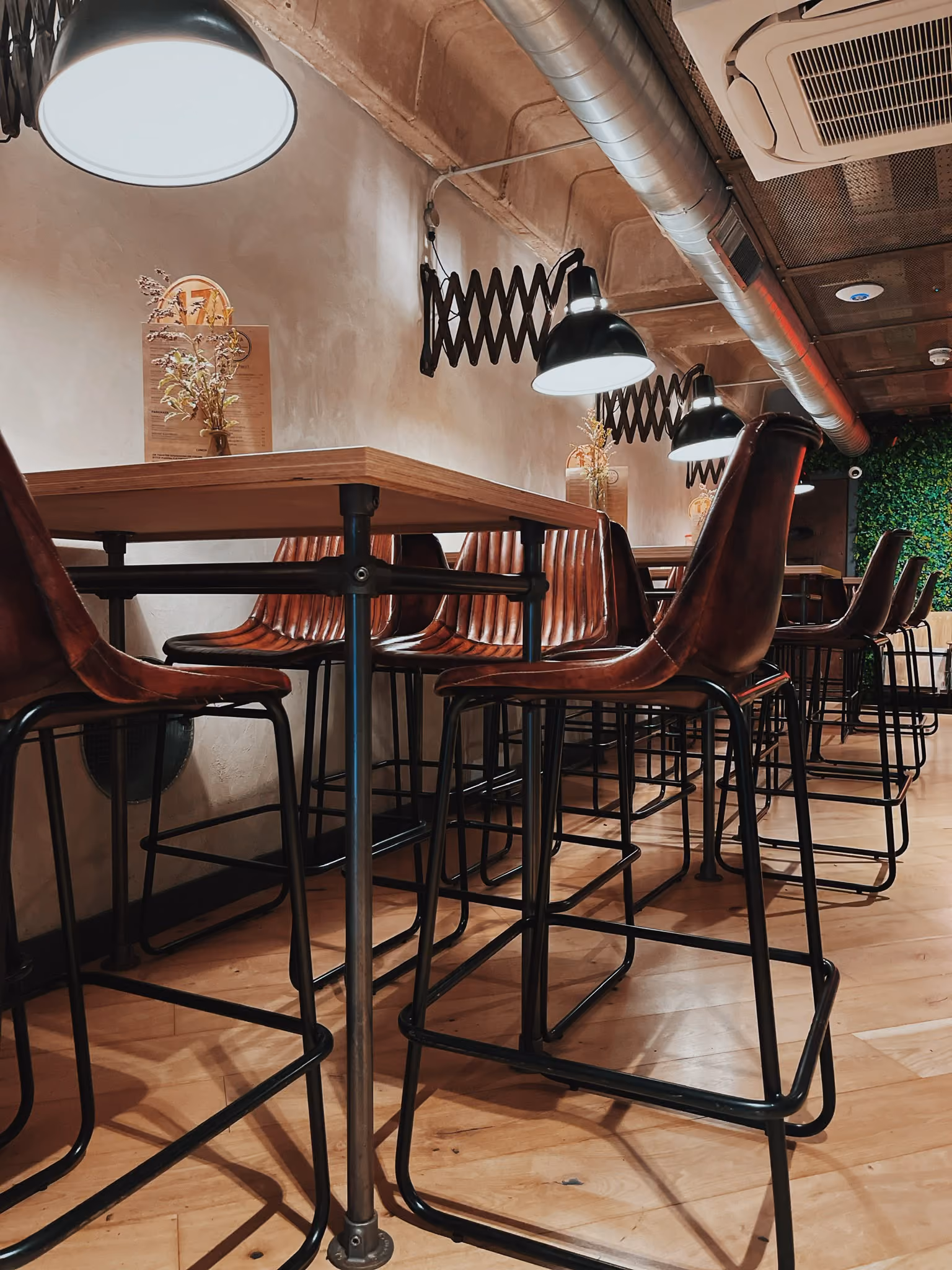 Rows of leather high stools at long tables under scissor-arm lamps at Ezra & Gil Peter Street