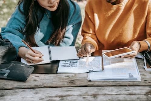 Zwei junge Frauen sitzen an einem Holztisch mit Bibel und Büchern, Symbol für Lernen und Austausch – Bild für die Kategorie Grundlagen im christlichen Blog.