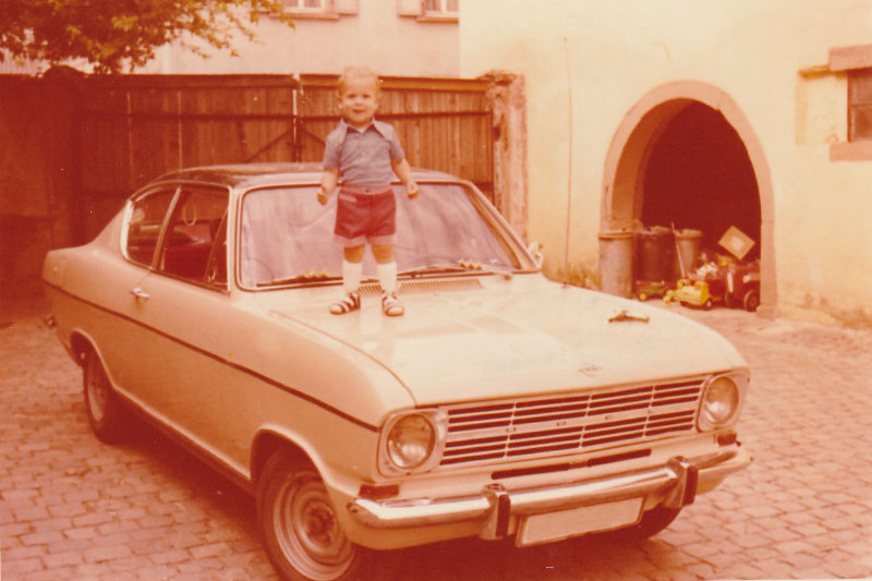 Dominik Bierle als Kind mit etwa 3 Jahren steht lachend auf der Motorhaube eines beigen Opel-Kadett im Hof seines Elternhauses – Beginn seiner Life-Story Wie werde ich glücklich.