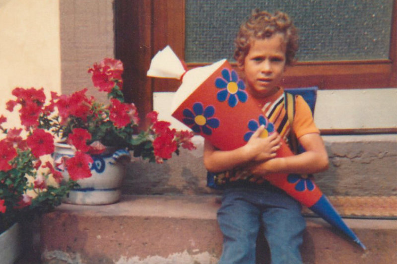 Dominik Bierle als kleiner Junge sitzt auf der Sandsteintreppe seines Elternhauses und hält eine sehr große, mit blauen Blumen dekorierte Schultüte, trägt lockiges Haar, blaue Jeans und einen blauen Schulranzen – ein Moment voller Kindheitserinnerungen und Lebensfreude.