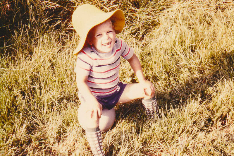 Der kleine Dominik Bierle mit etwa 3 bis 4 Jahren sitzt lachend auf einem weißen Fußball im Gras, trägt einen gelben Schlapphut, kurze Hosen und lange Socken – ein fröhlicher Moment seiner Kindheit, passend zur Life-Story Wie werde ich glücklich.