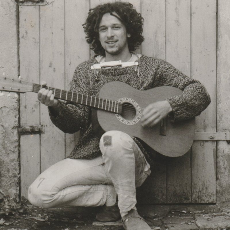 Dominik Bierle mit Dreadlocks, Mundharmonika und Gitarre im Bob-Marley-Stil, vor einer alten Holztür mit selbstgestricktem Pullover – ein Symbol für die Suche nach dem Sinn des Lebens und nach Freiheit.