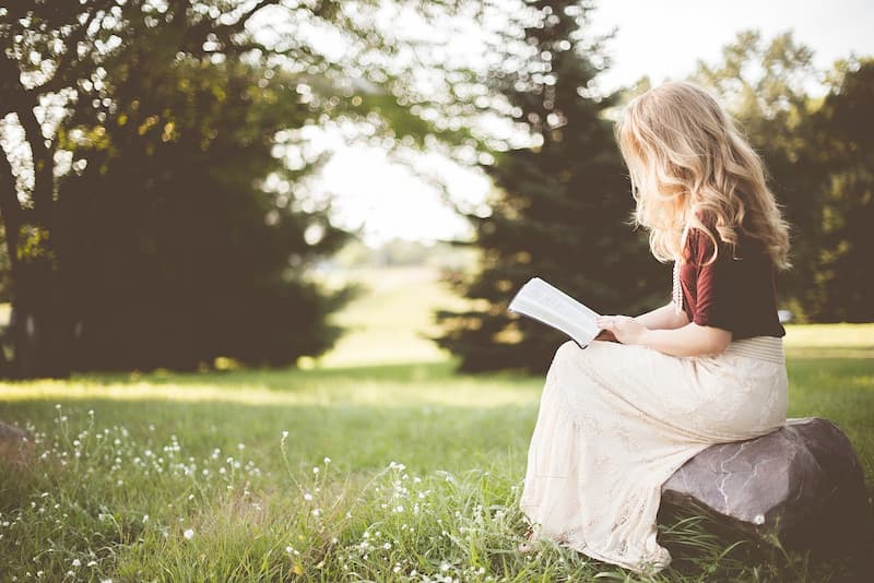 Frau sitzt auf einem großen Stein auf einer grünen Wiese, Beine überschlagen, liest die Bibel, trägt einen weißen langen Rock und hat lange blonde Haare – Symbol für Ruhe, Andacht und persönliche Bibellektüre.