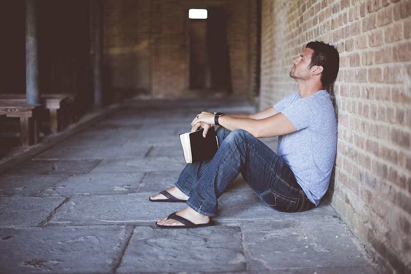 Mann sitzt auf einem Steinboden in einer Kirche, lehnt mit dem Rücken an eine Steinmauer, Augen geschlossen, denkt über die Bibel nach und hält seine Bibel in der Hand – Symbol für Andacht, Reflexion und persönliche Bibellesung.