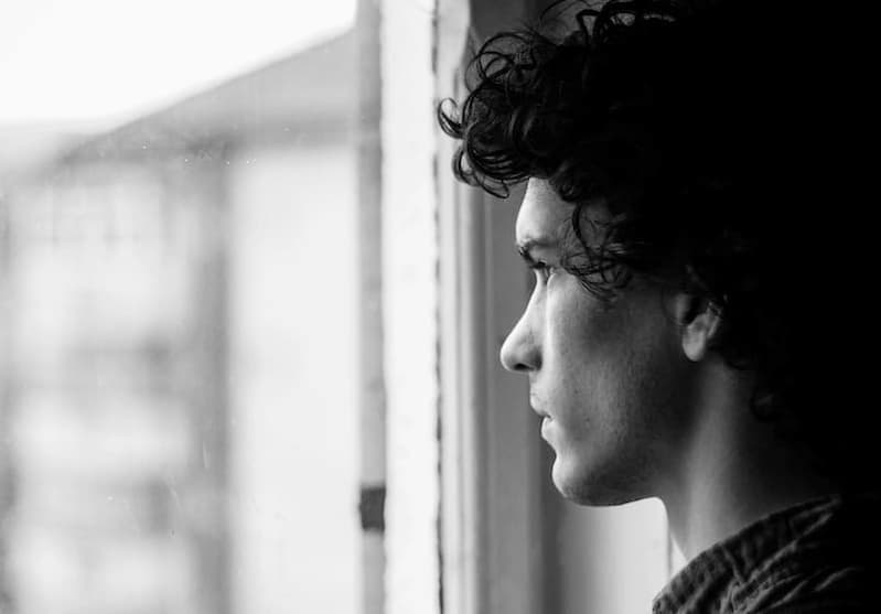 Nachdenklicher junger Mann mit schwarzen Locken schaut aus dem Fenster und fragt sich: Wer ist Gott? Diese Szene symbolisiert die Suche nach Gott, das Fragen nach Gottes Wesen und die Sehnsucht nach Antworten.