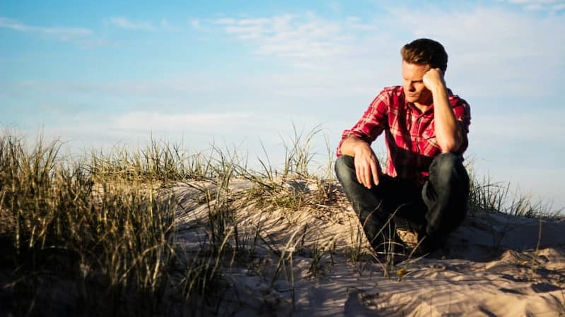 Mann sitzt nachdenklich auf einer Düne am Strand und überlegt, welche schwierige Entscheidung er treffen soll