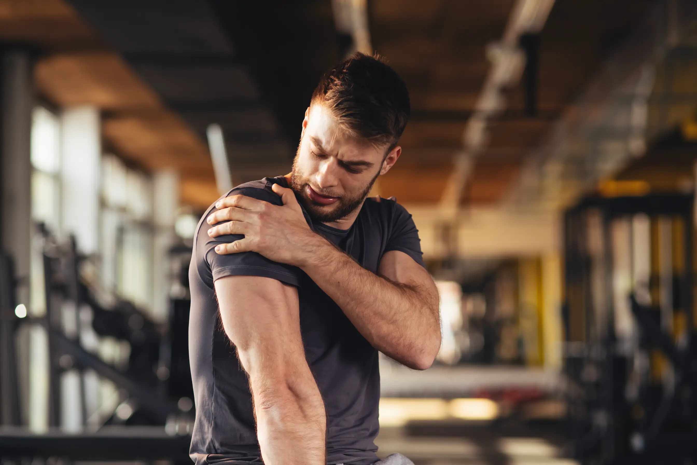 Young man in a gym gripping his shoulder with a pained expression, hinting at a sports injury or muscular discomfort, showed here as a great example of the patient who would do well with shockwave therapy. 