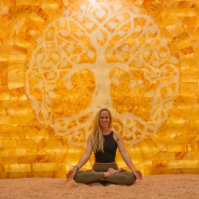 Woman sitting on a salt covered floor smiling in front of a LED backlit salt panel with a shape of a tree within at the Purify Wellness Center - Pleasant Grove, UT.